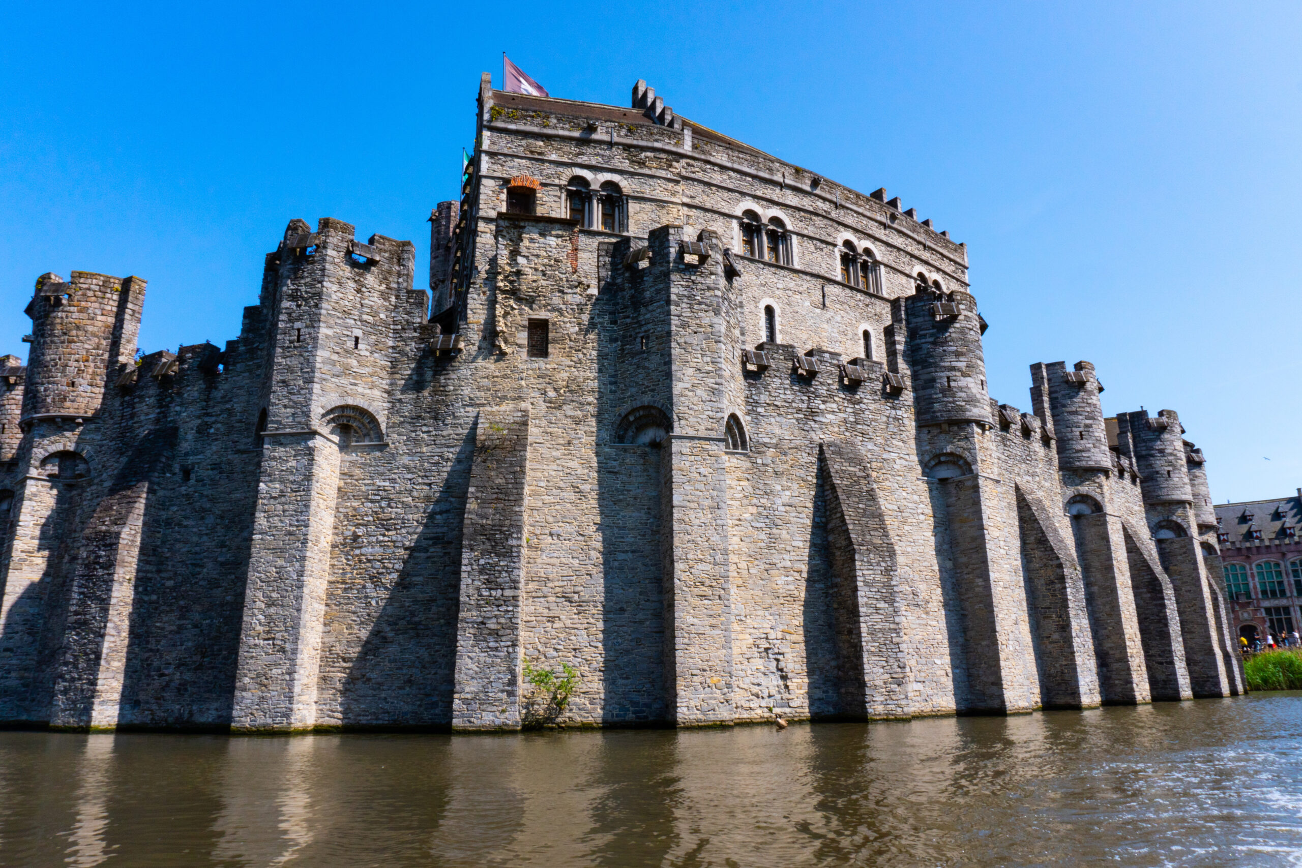 Stone medieval fortress with tall towers and thick crenellated walls along a river under a blue sky