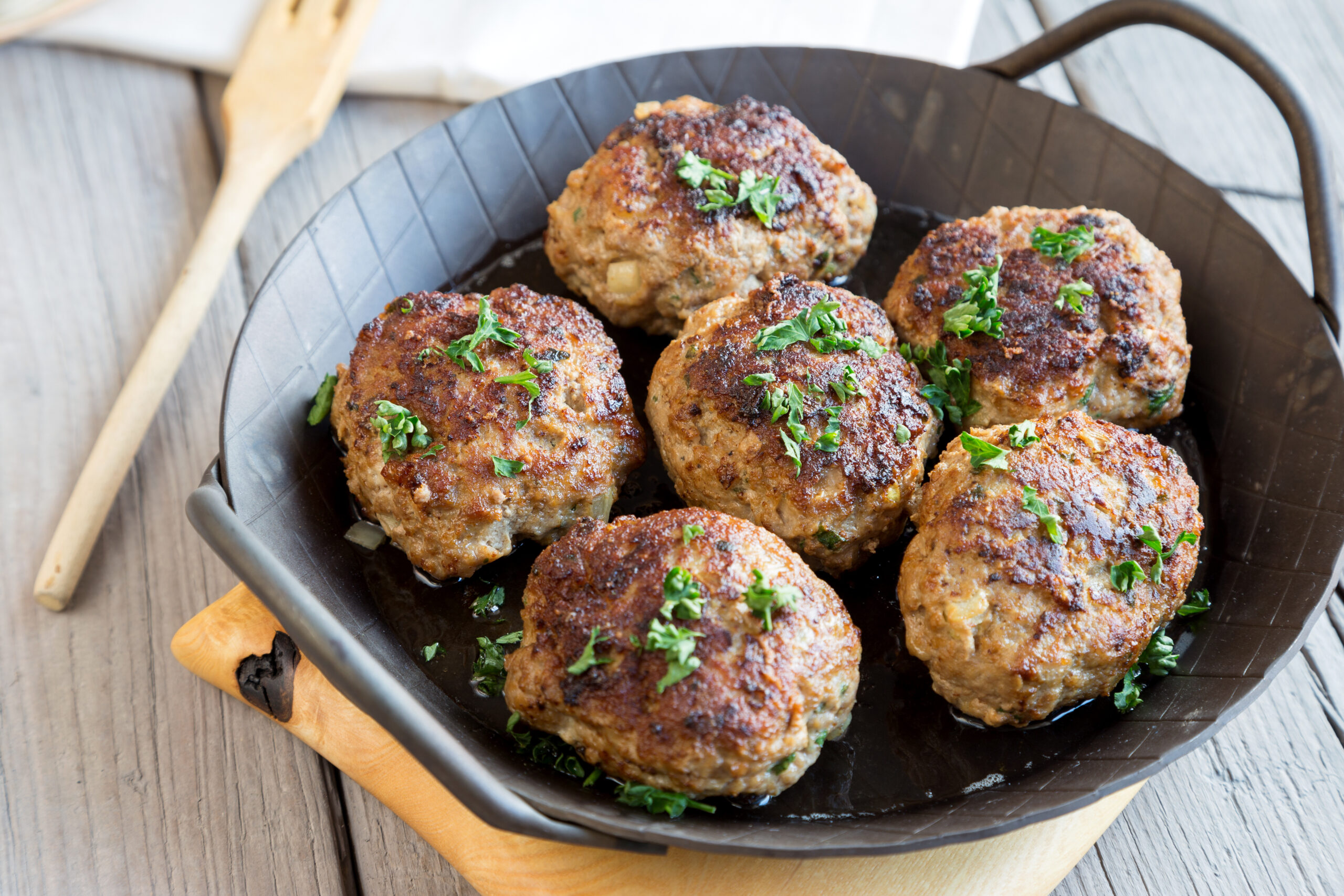 Browned meatballs in a black skillet, sprinkled with chopped parsley on a wooden table.