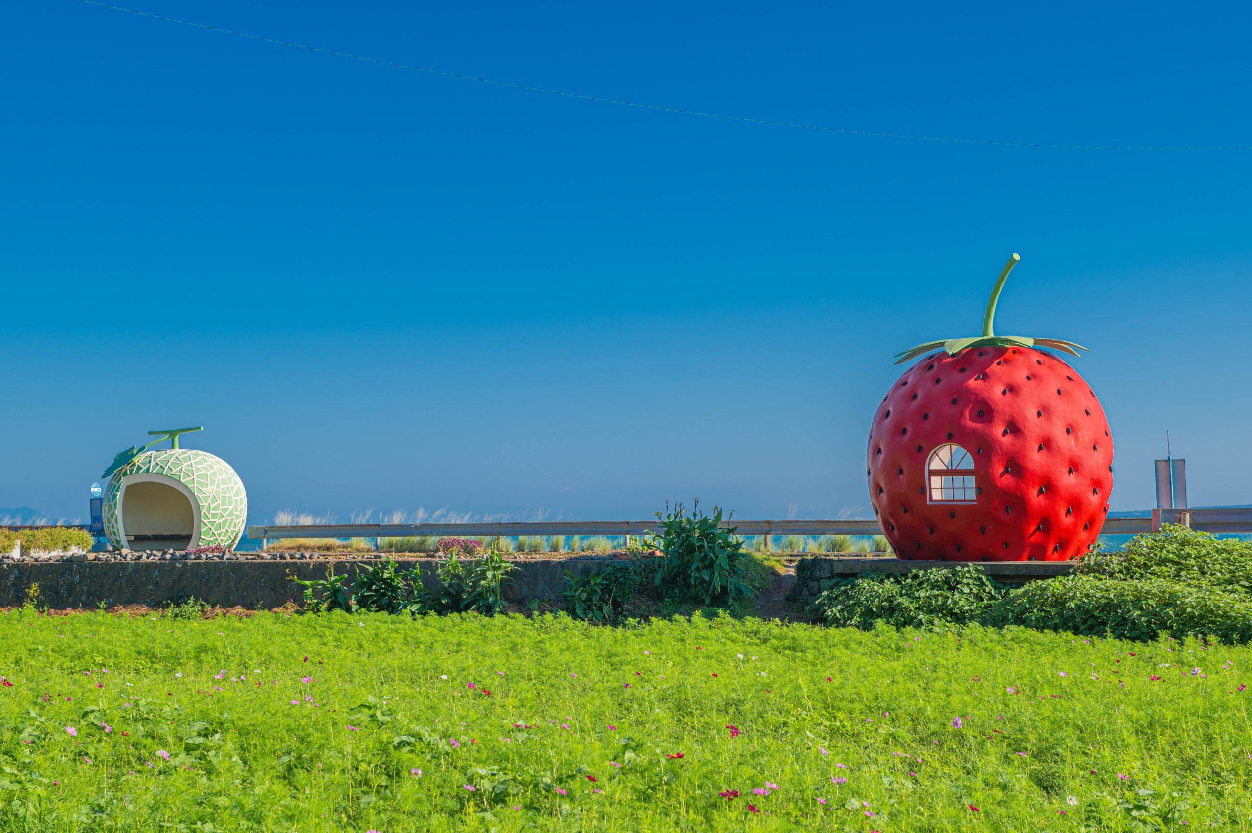 Giant red strawberry-shaped house with window and green stem in a sunny field under a blue sky