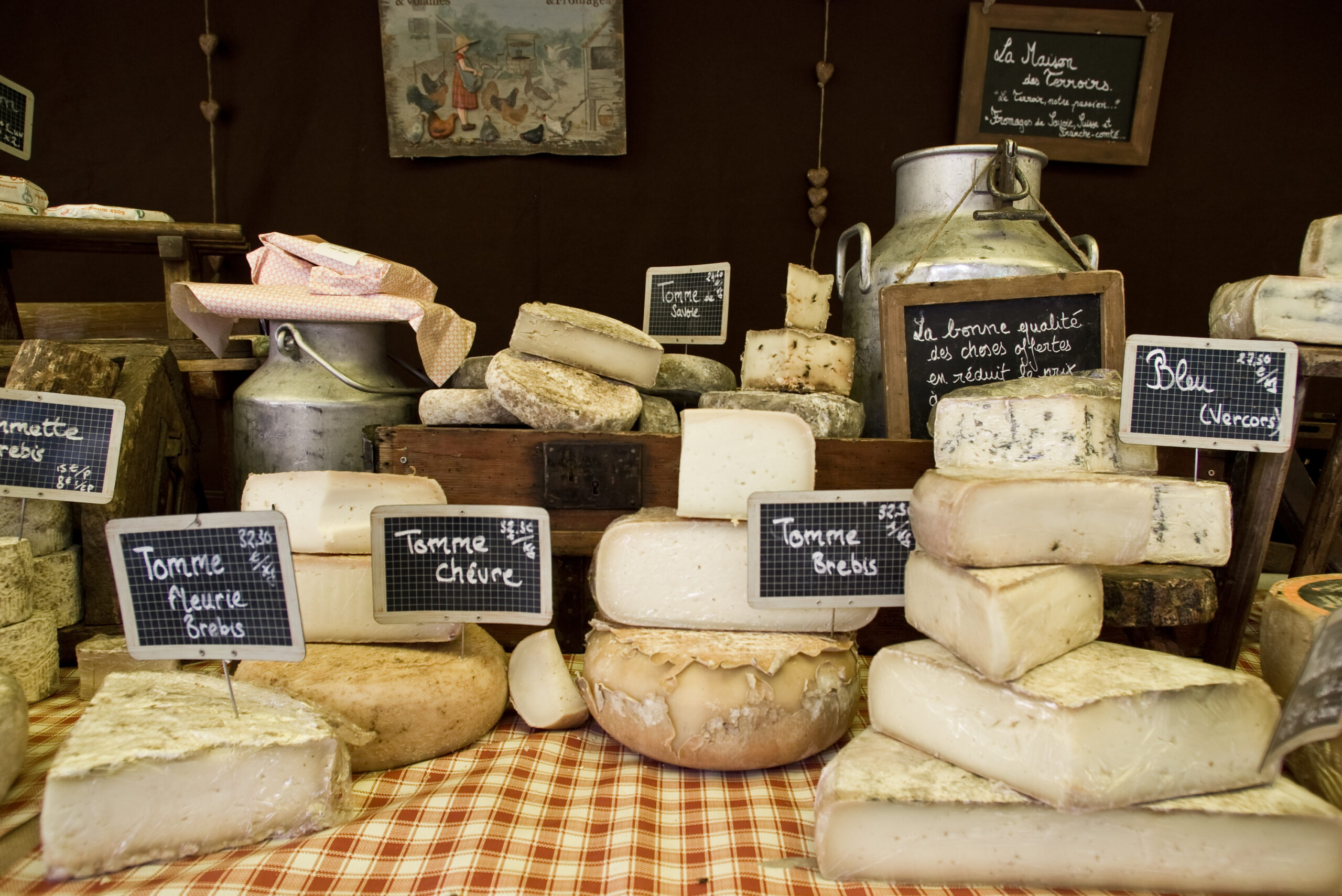 Assorted cheeses stacked on a checkered cloth with small chalkboard signs listing types such as Tomme chèvre and other varieties.