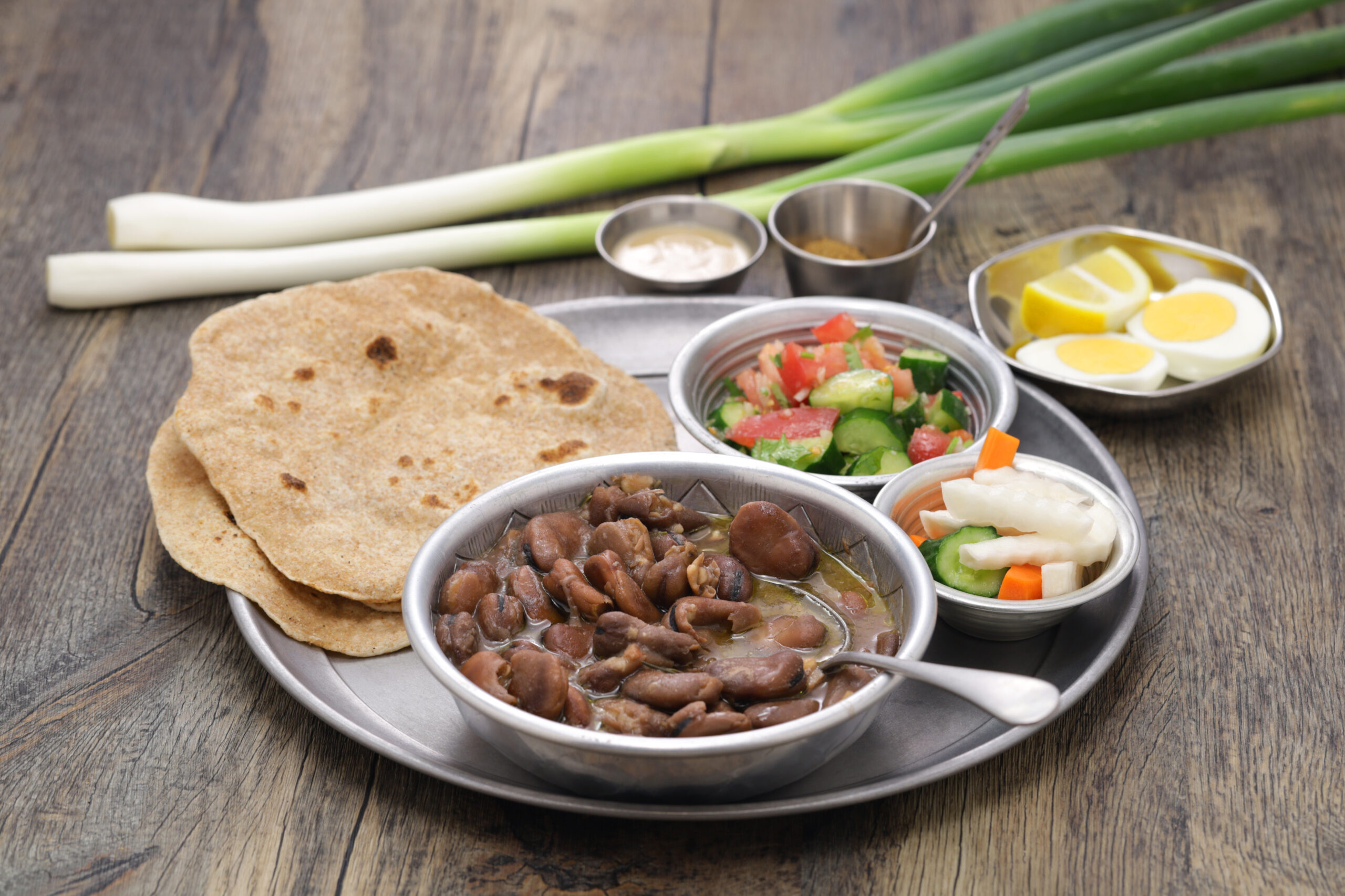 Metal platter with tortillas, refried beans, pico de gallo, pickled vegetables, and boiled egg halves with lemon wedges on a rustic wooden table, plus green onions in the background.