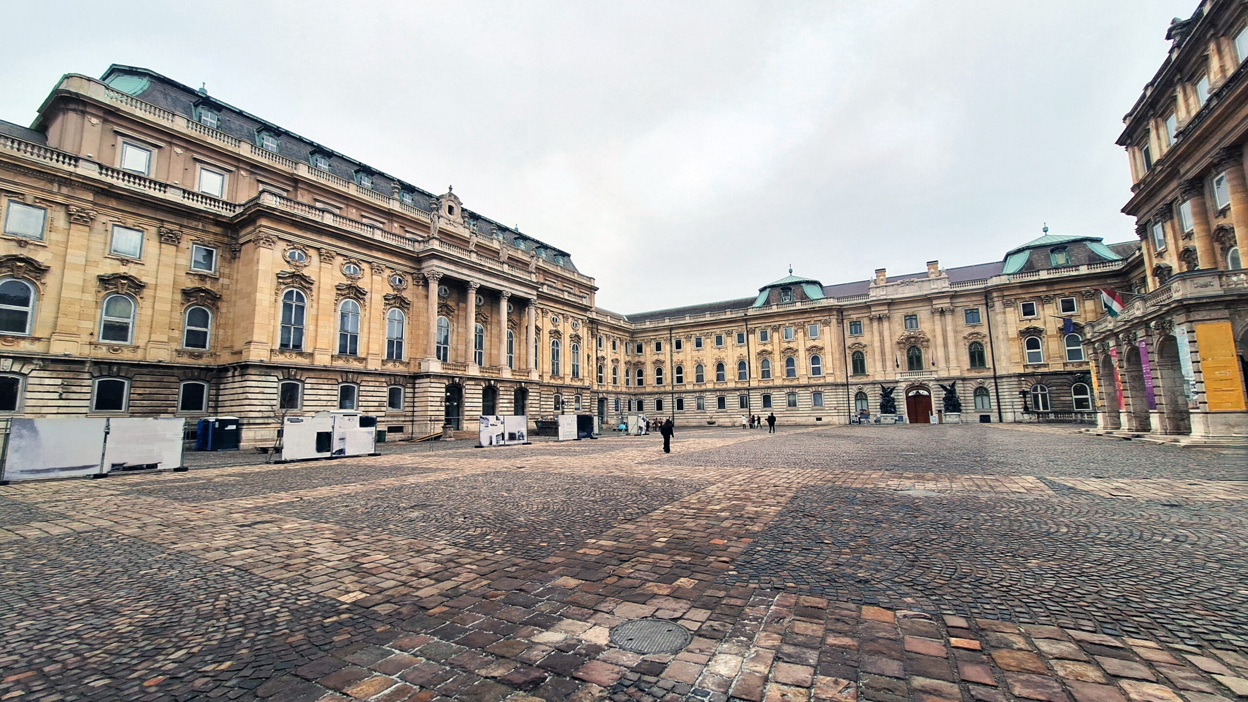 Wide view of a classical beige stone building complex surrounding a cobblestone plaza, with arched windows and decorative columns