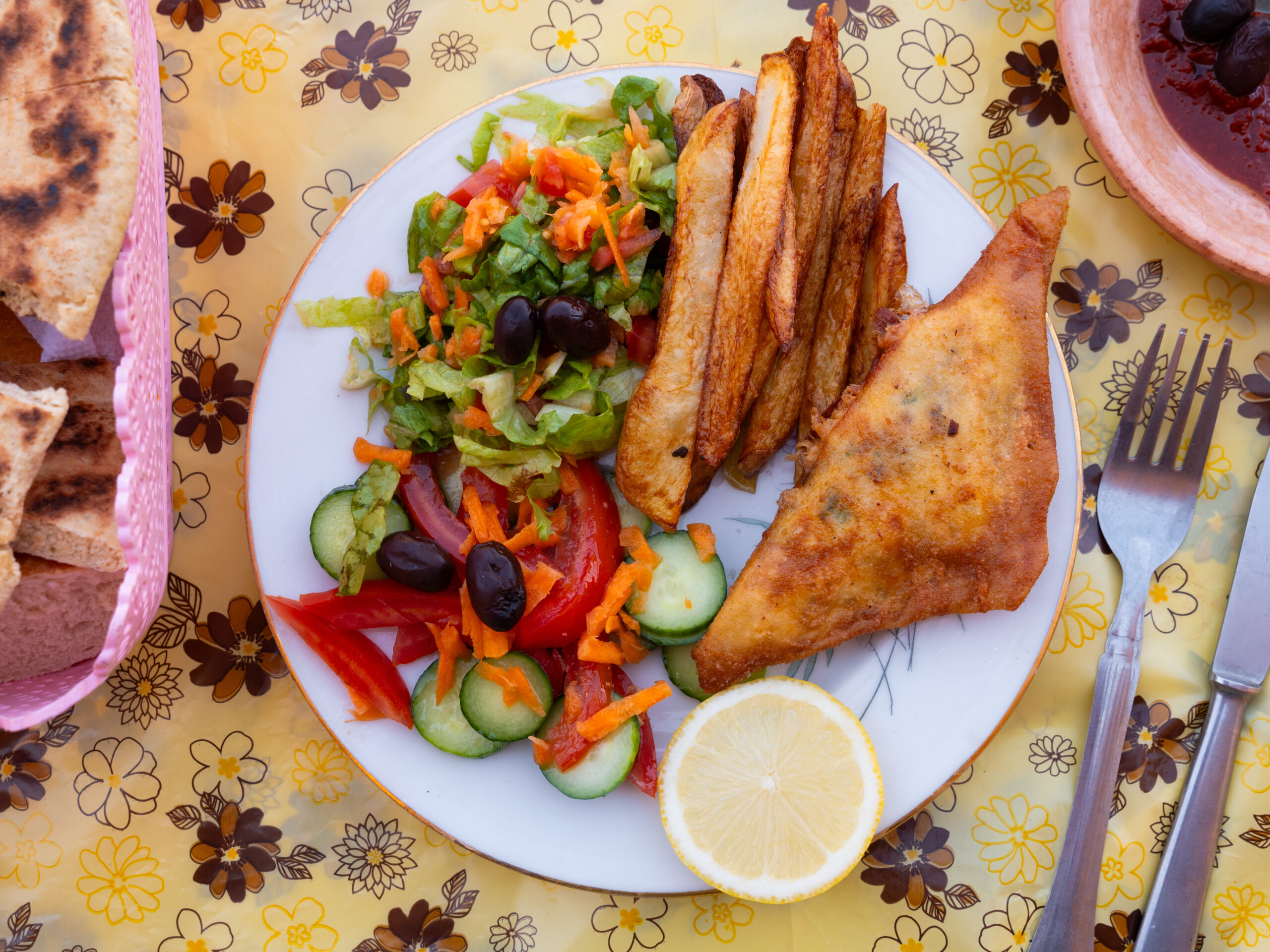Plate with mixed salad (lettuce, cucumber, tomatoes, olives, shredded carrots) and several fried plantains, plus lemon wedges on a tablecloth.