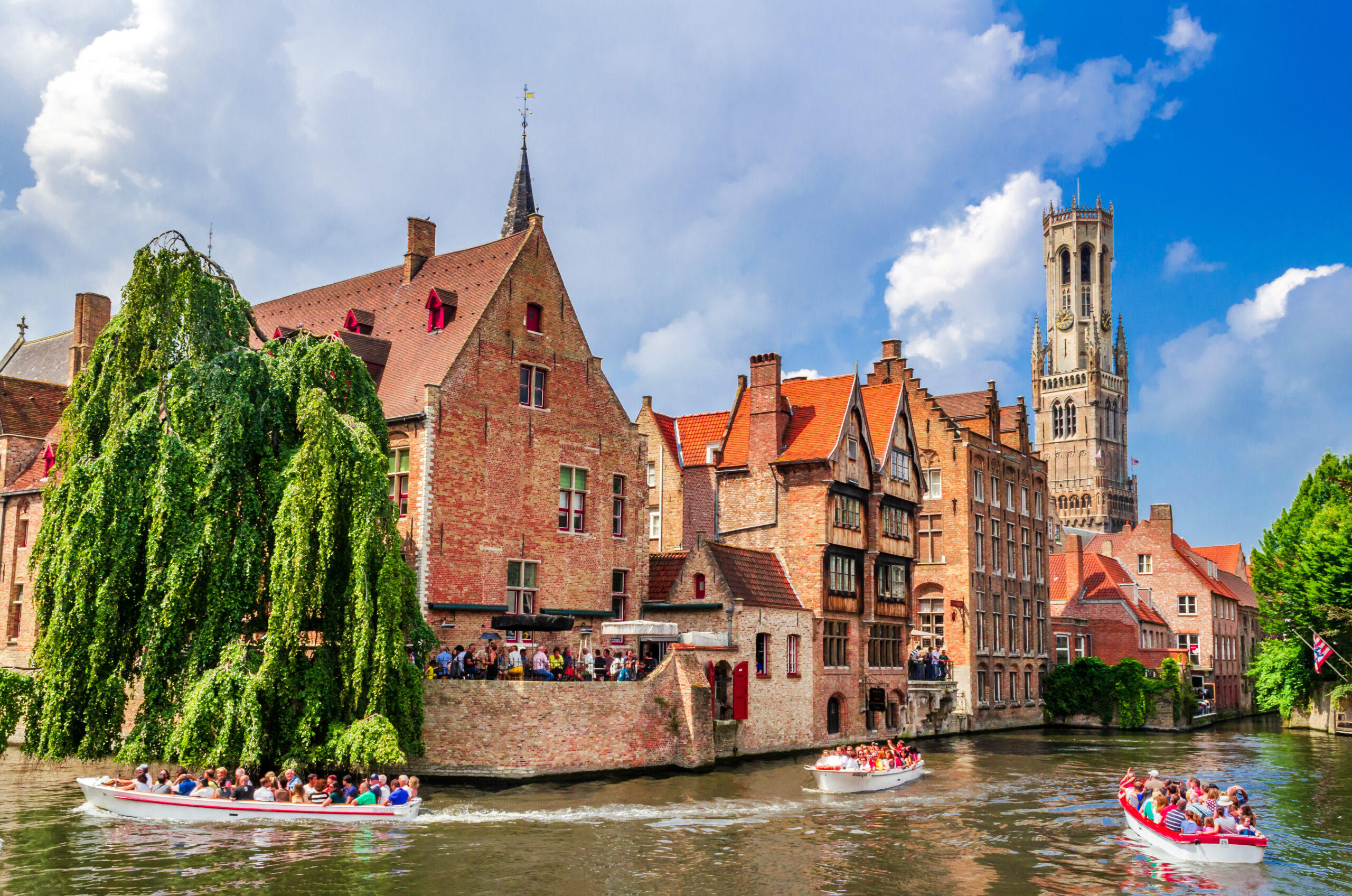 Tour boats carrying people glide along a canal beside red-brick medieval buildings in Bruges, with a tall church tower in the background under a blue sky.”
