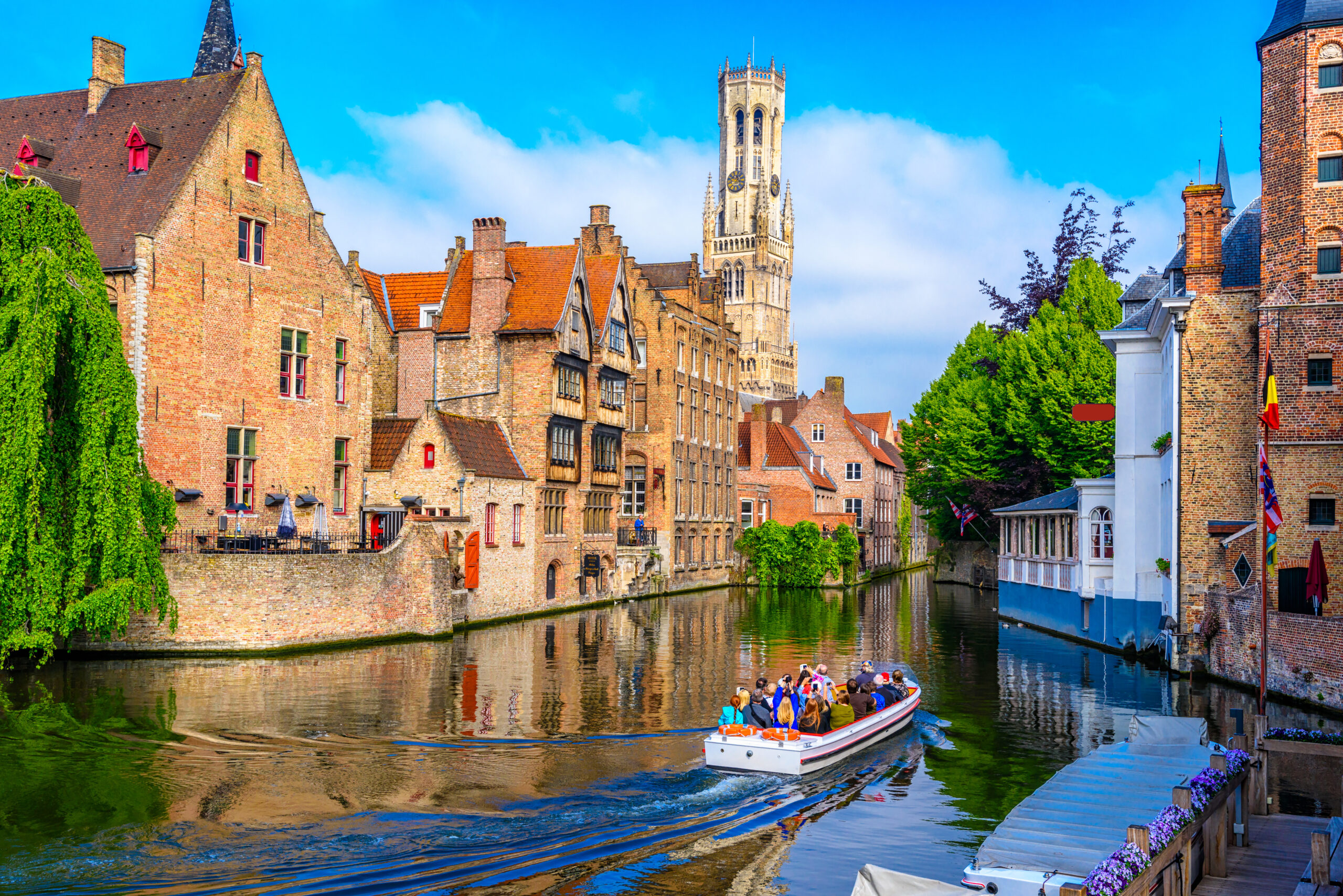 Canal scene with historic brick buildings along the water and a tall church tower in the distance; a tour boat with passengers glides by.