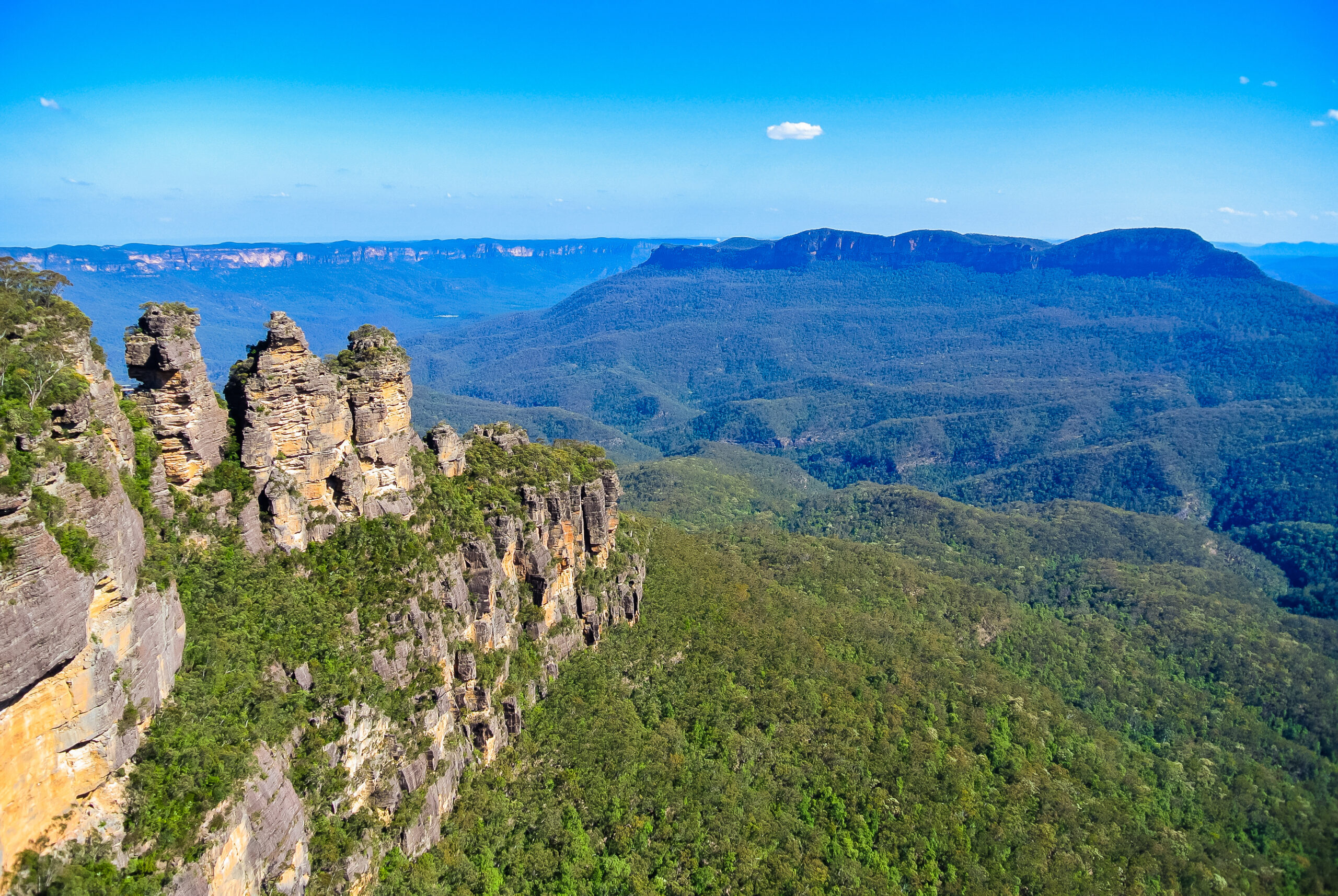 Rocky cliff spires on the left with a dense forest below and distant blue mountains under a clear sky, wide landscape view.