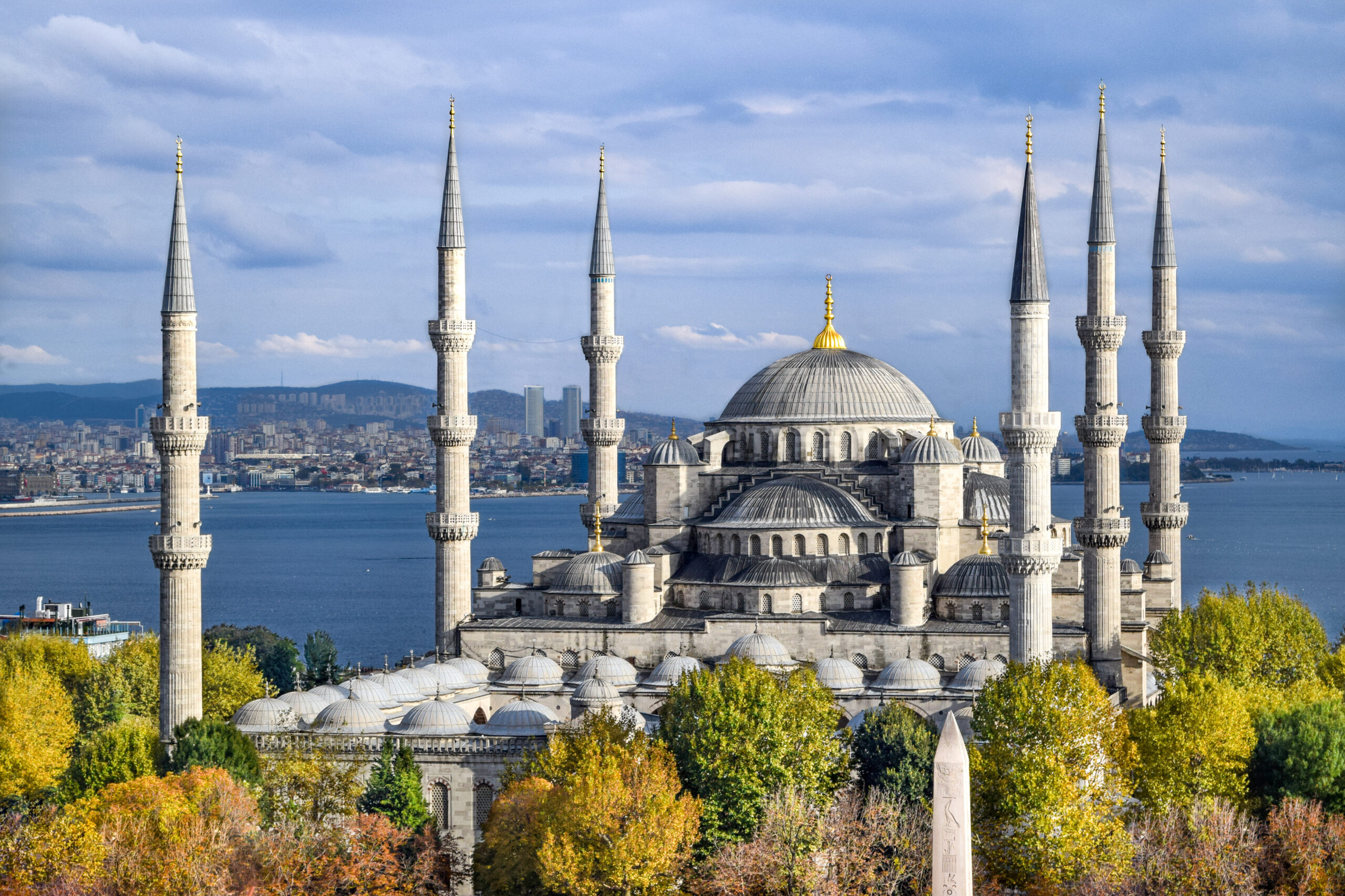 Blue Mosque in Istanbul with six minarets and a large central dome, framed by autumn trees and a water backdrop