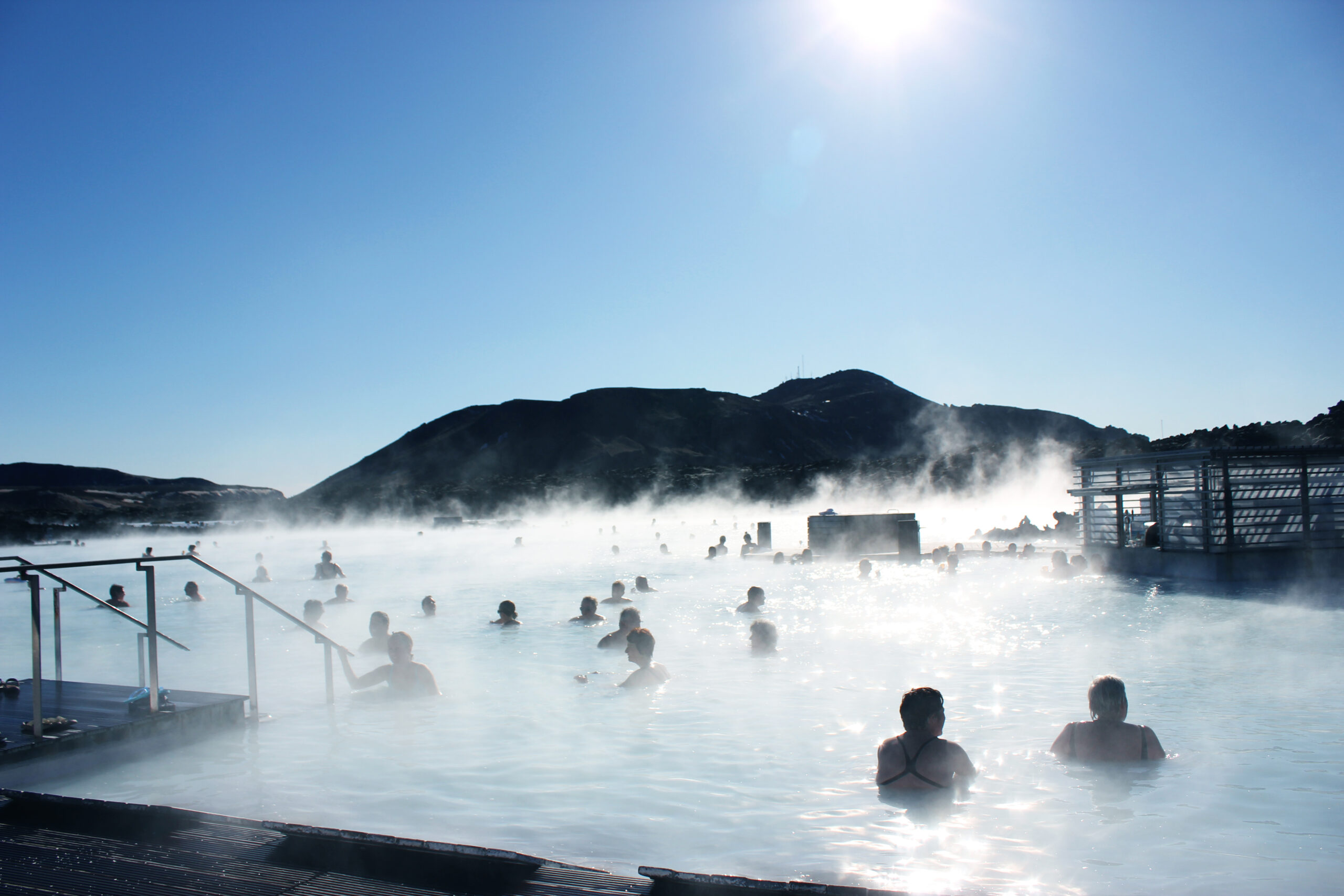 People soak in a steaming outdoor hot spring with a mountain backdrop under a bright blue sky.