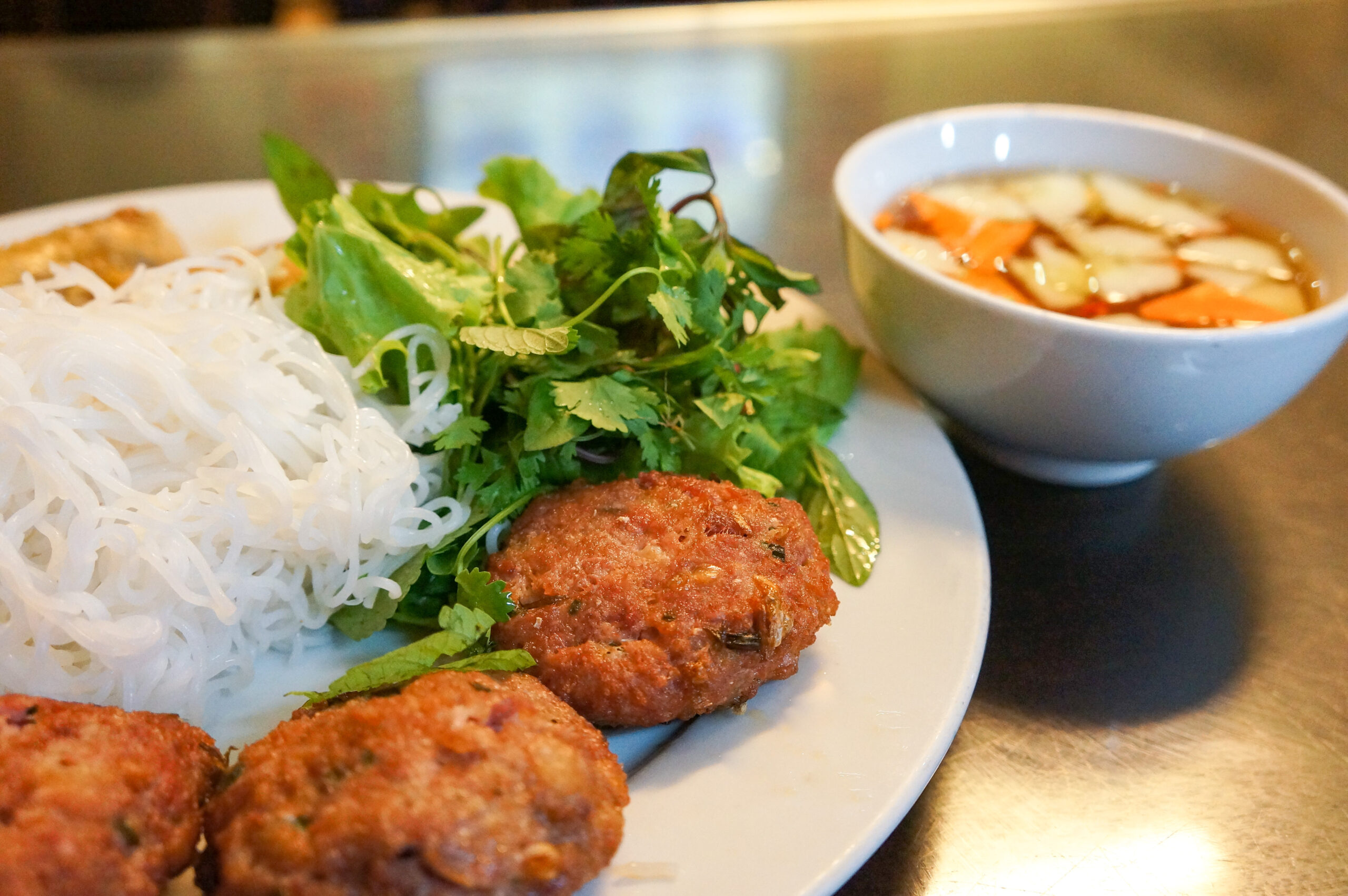 Plate with white rice noodles, fresh herbs, and fried patties; a bowl of soup in the background.