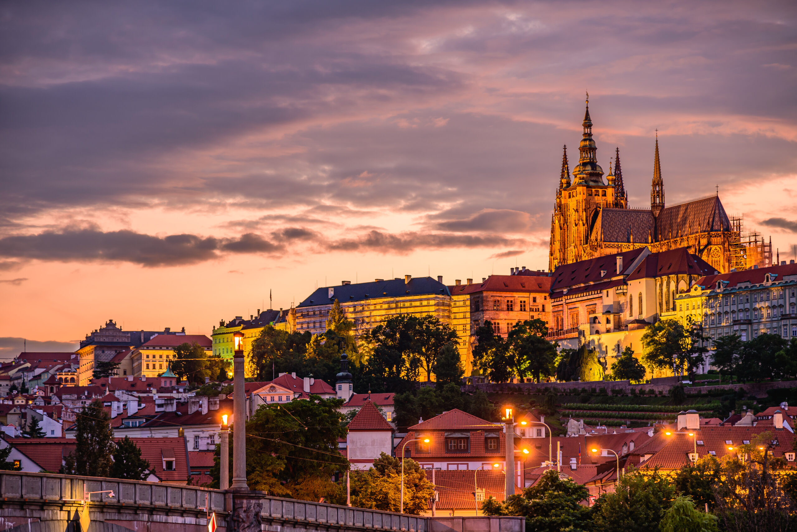 Sunset over a European city with a Gothic cathedral and colorful rooftops, warm light bathing the stone spires.