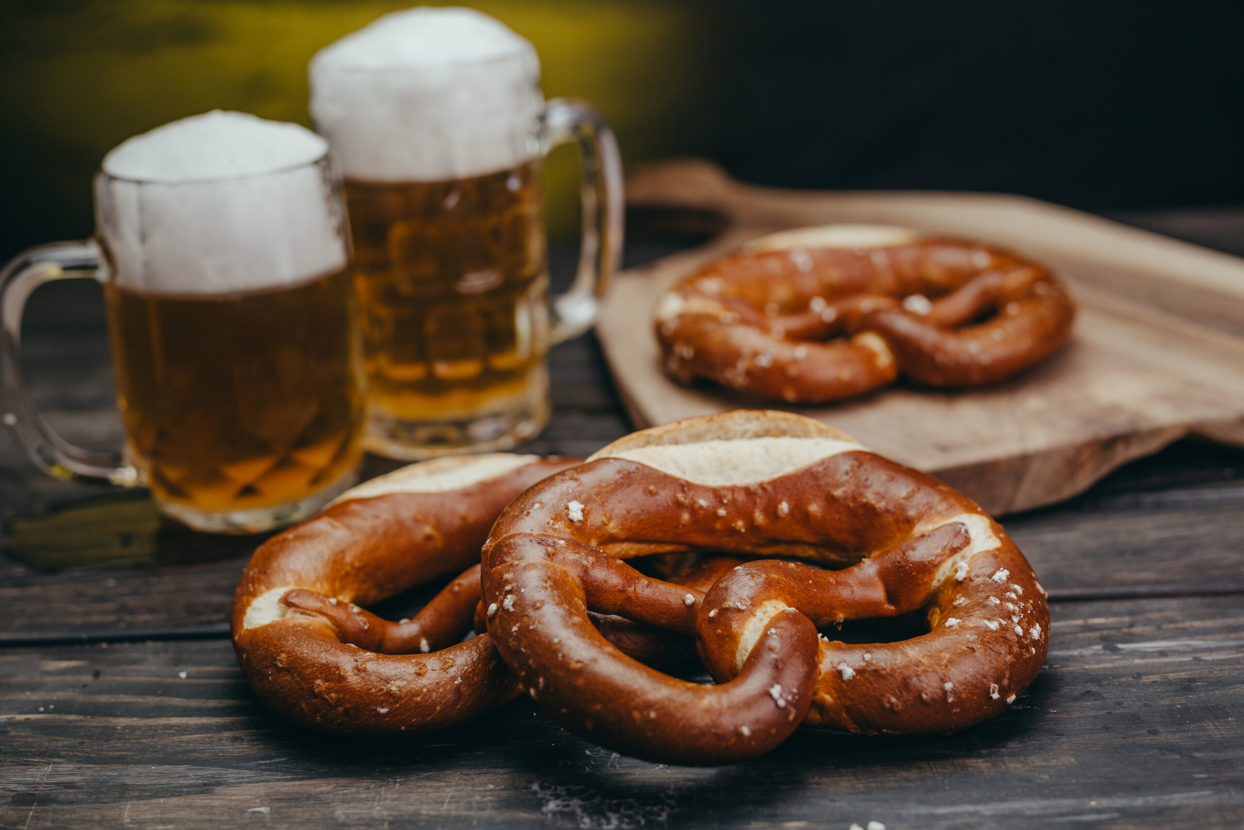 Two beer mugs with foamy heads sit beside a plate of salted pretzels on a wooden table.