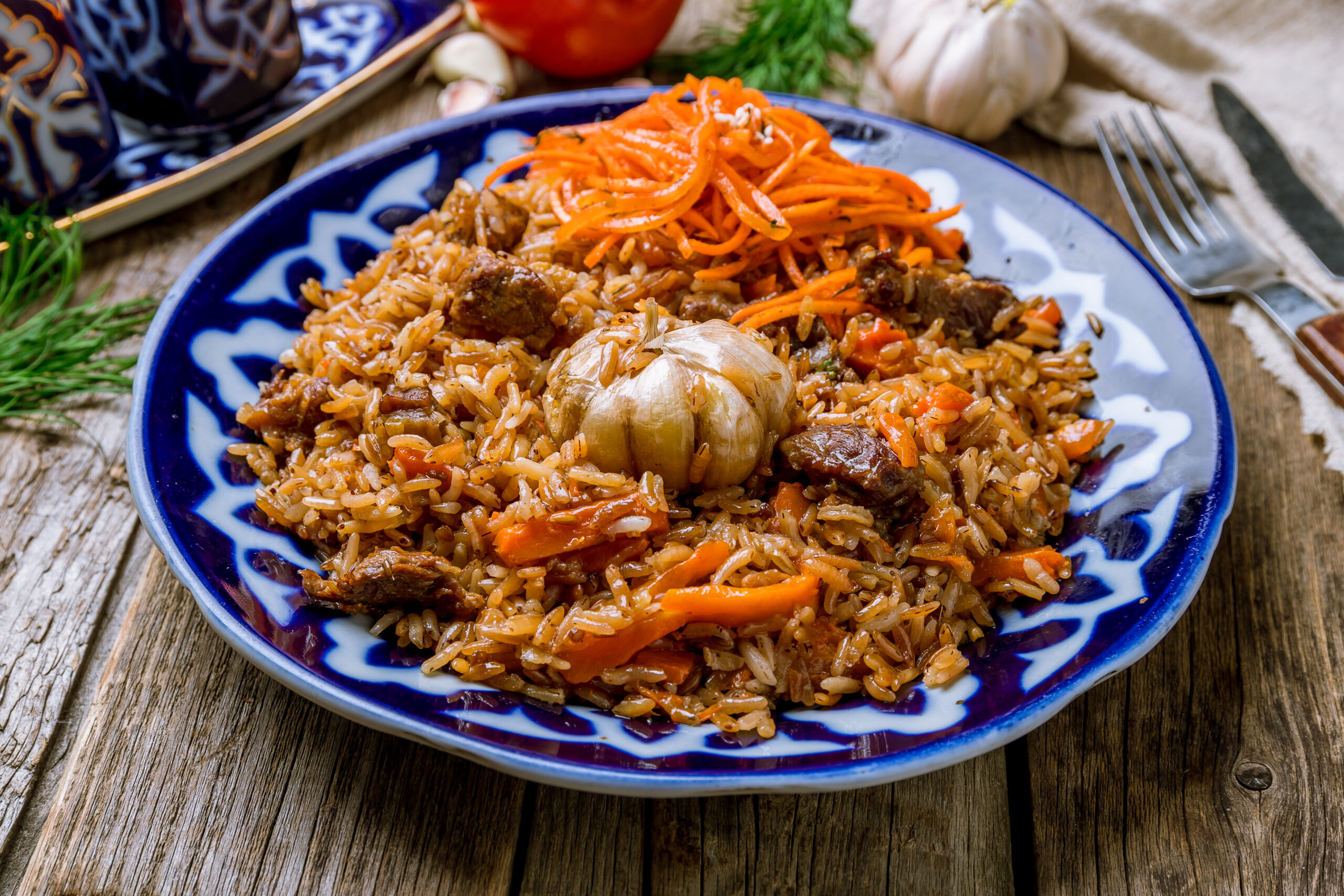 Plate of seasoned rice with beef chunks and carrot slivers on a blue patterned dish, garlic head in foreground and rustic wooden table backdrop.