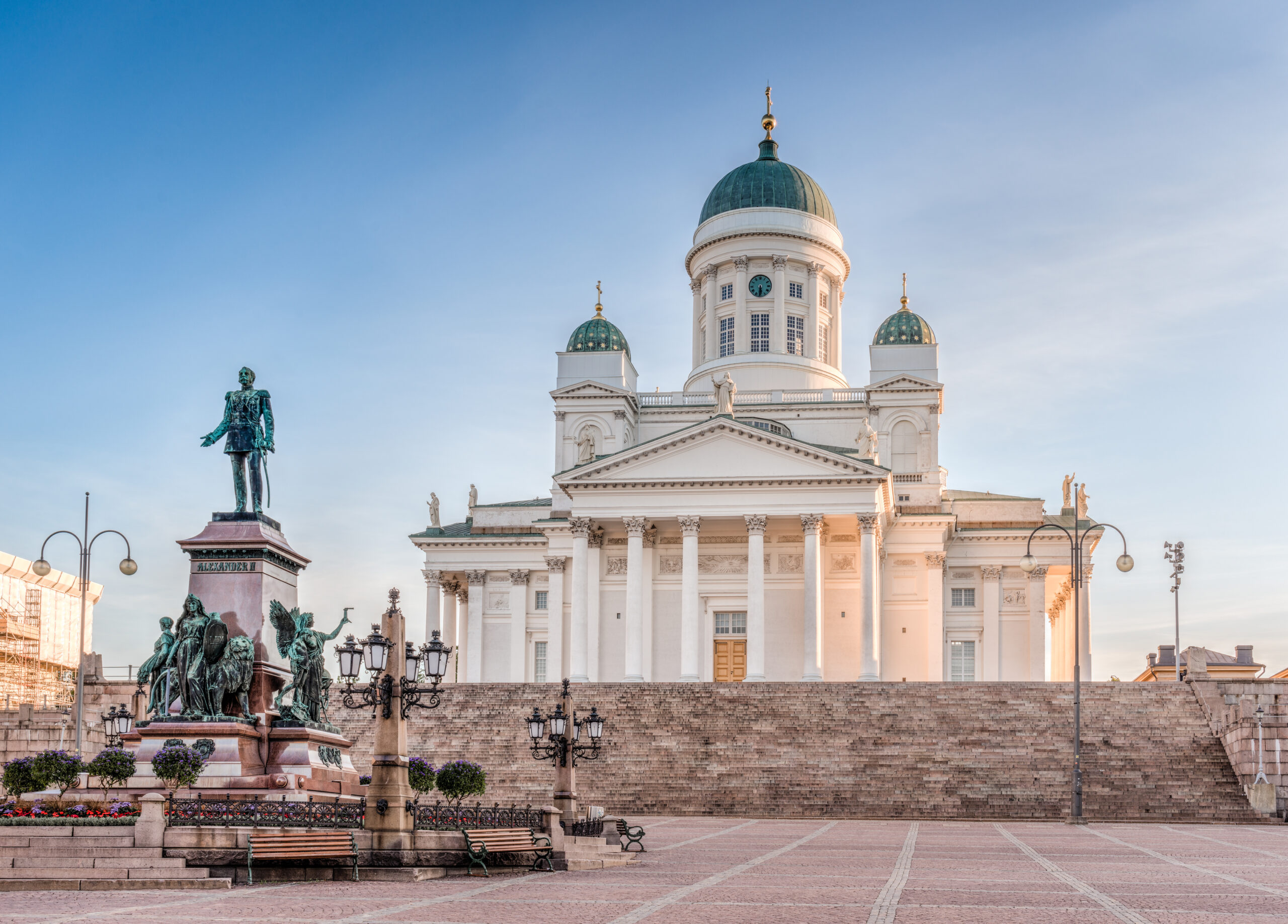 Neoclassical cathedral with three green domes and a clock, flanked by statues in a sunlit square in front of it.