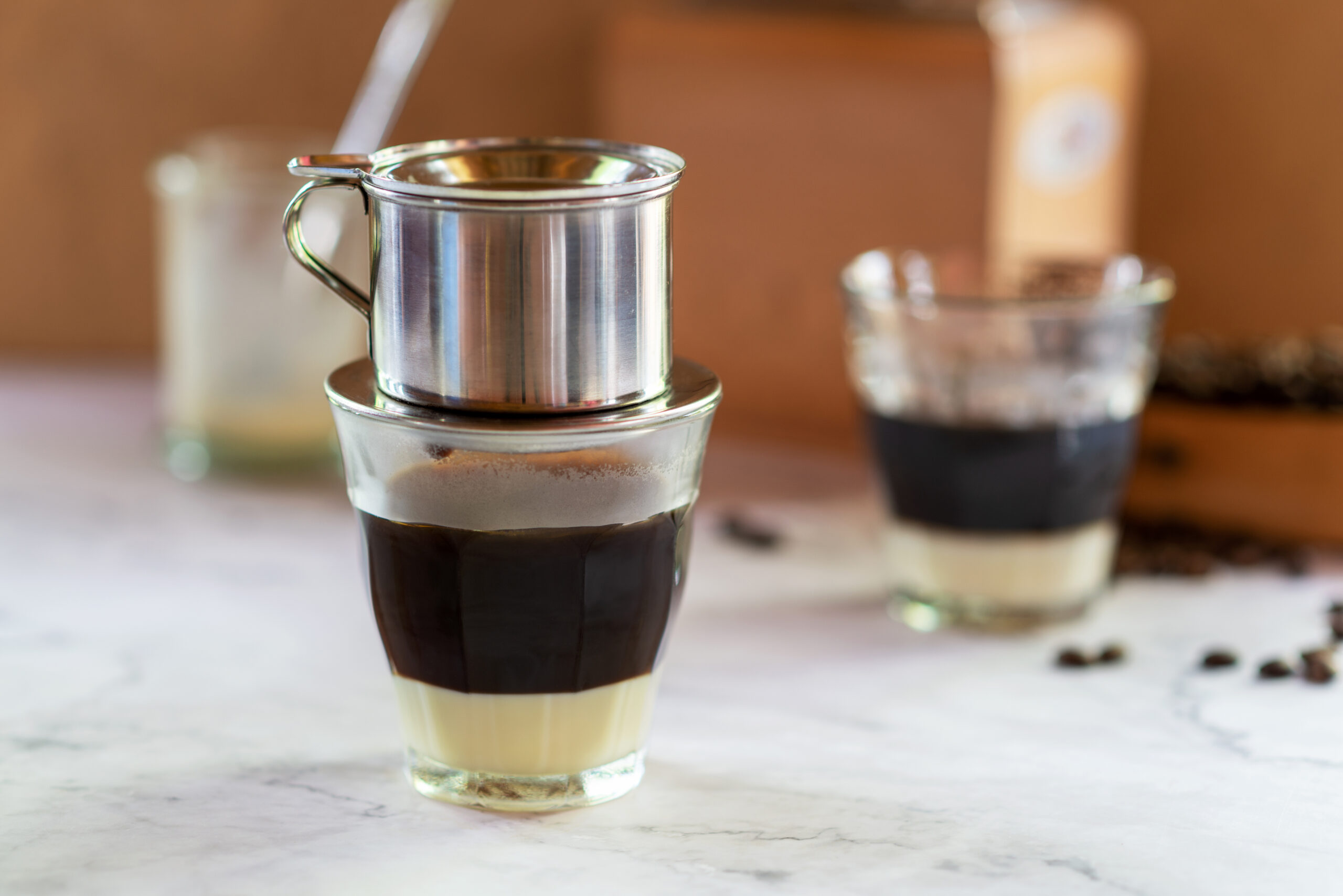 Layered espresso in a glass with a metal filter on top, blurred cups in the background and coffee beans to the right.