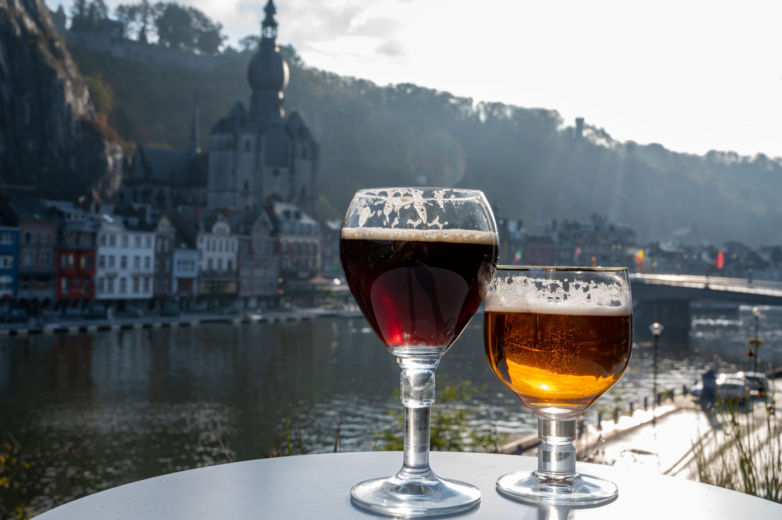 Two beer glasses on a white table with a riverside town and castle in the blurred background.