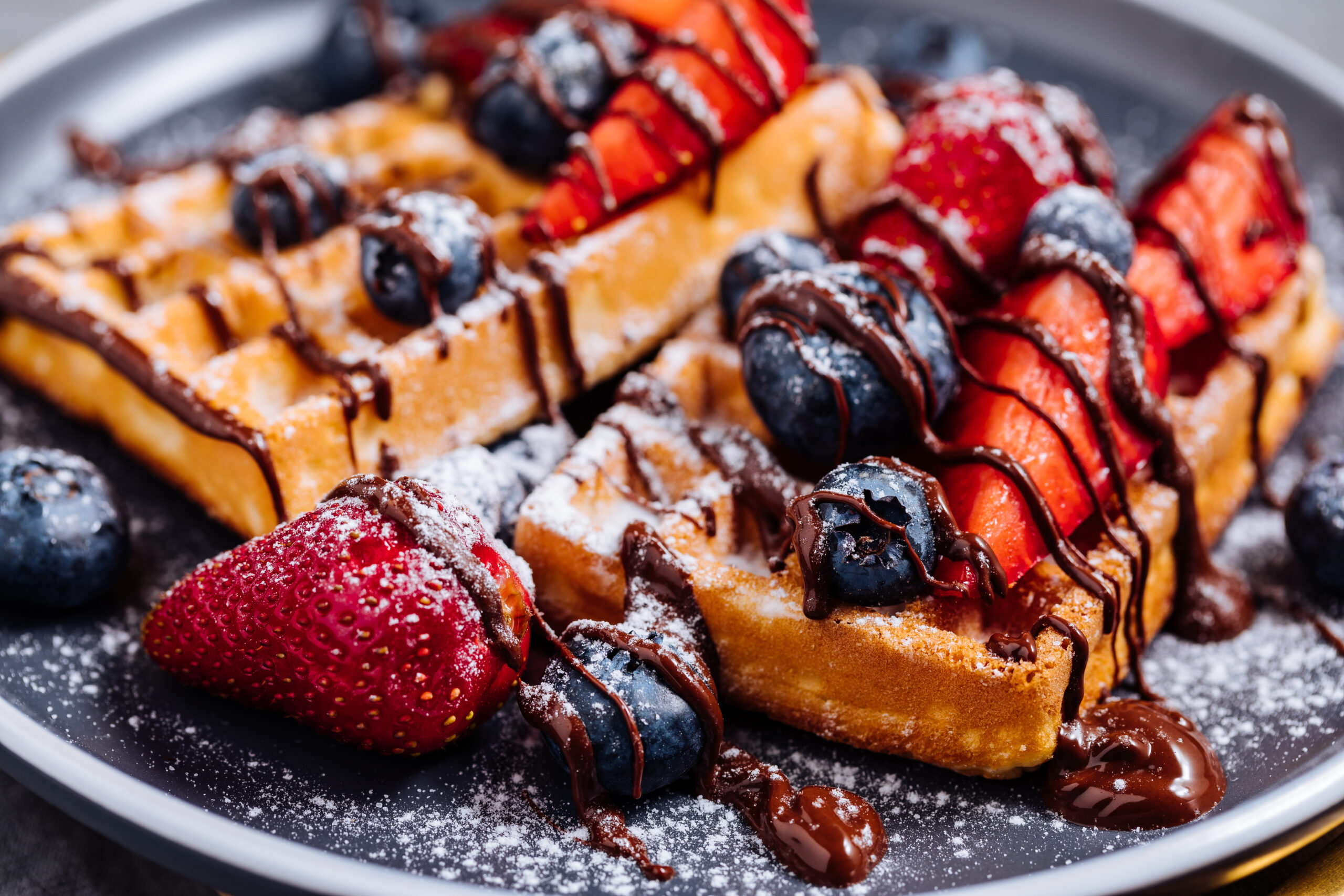 Plate of waffles topped with strawberries, blueberries, and chocolate drizzle, dusted with powdered sugar on a dark plate.
