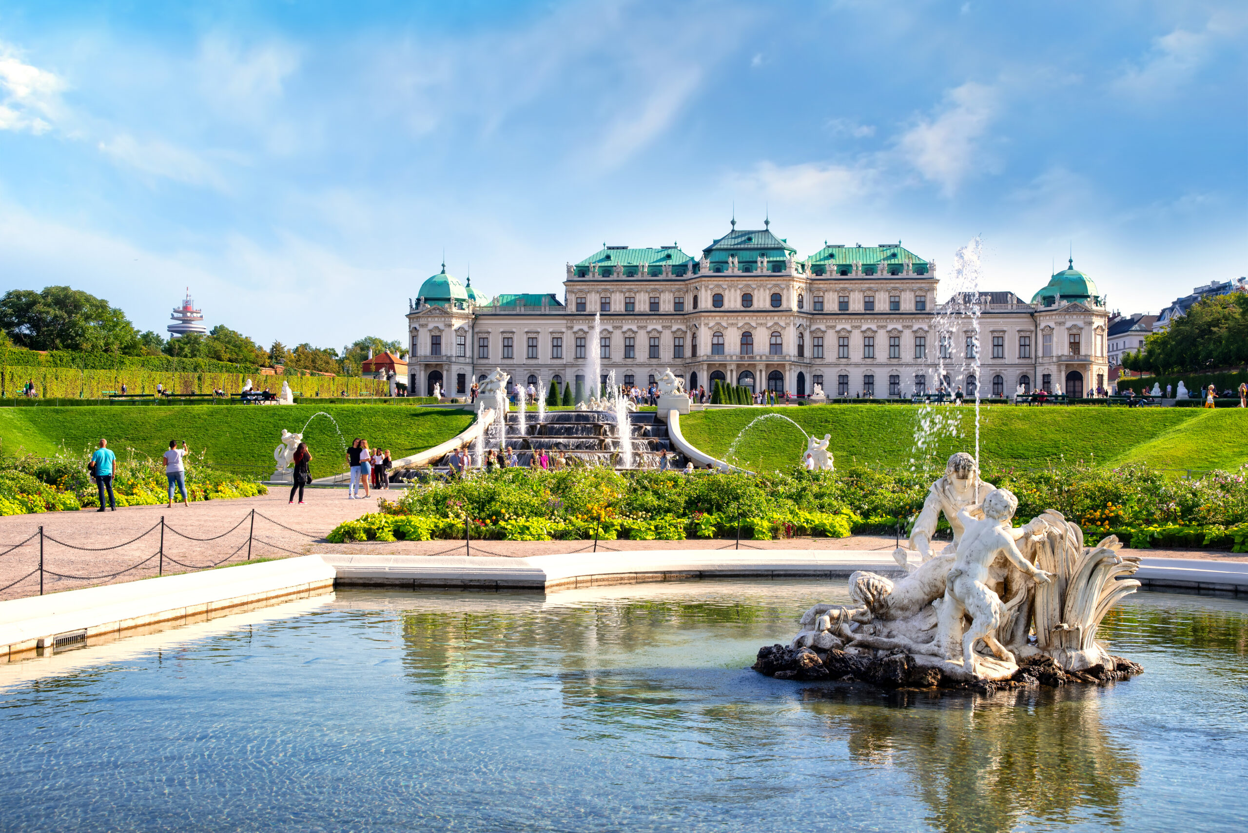 Grand white palatial building with green copper roofs, flanked by fountains and a manicured garden, under a blue sky with visitors nearby.