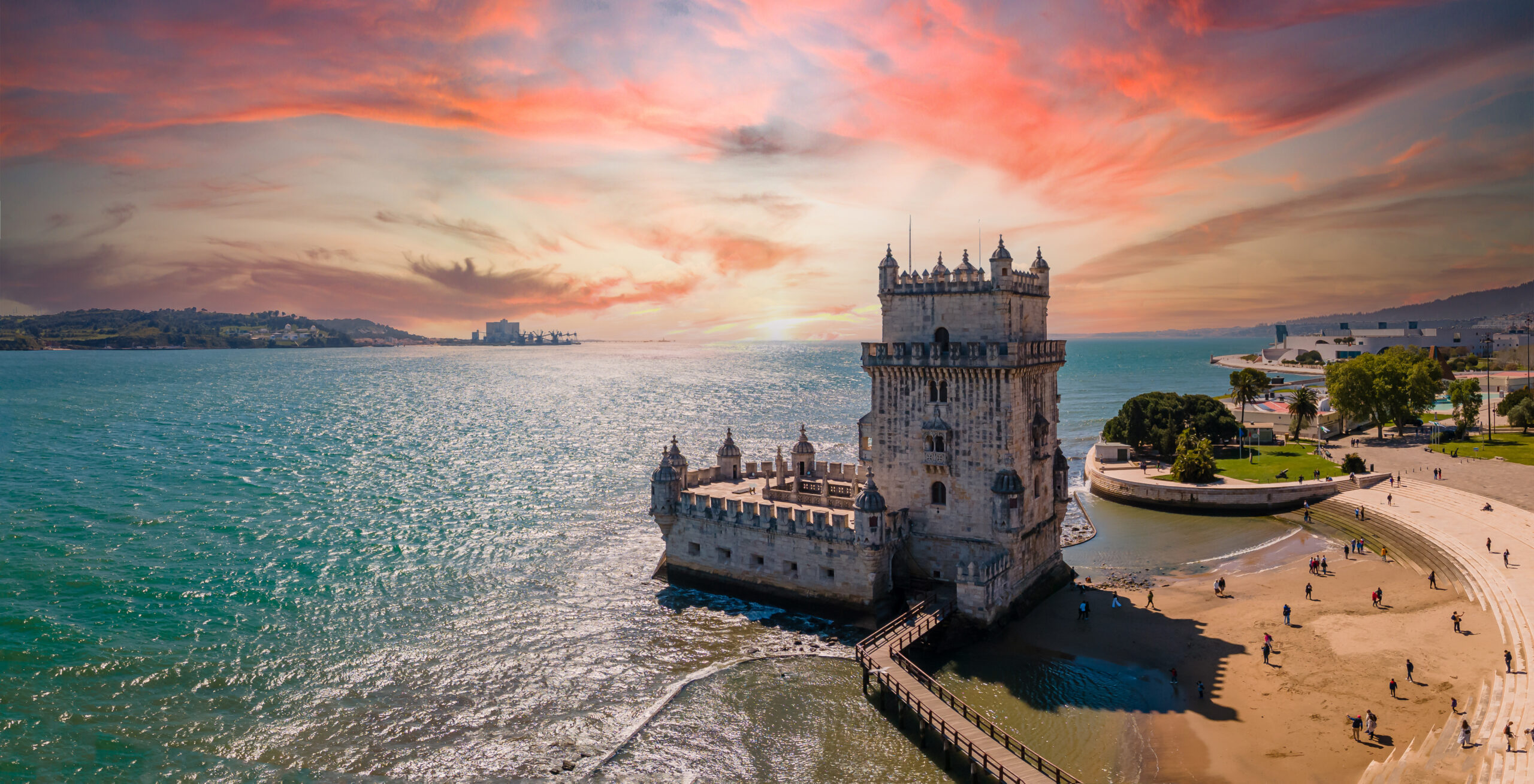 Belem Tower rising from the Tagus River at sunset, with a curved quay and people strolling along the waterfront park.