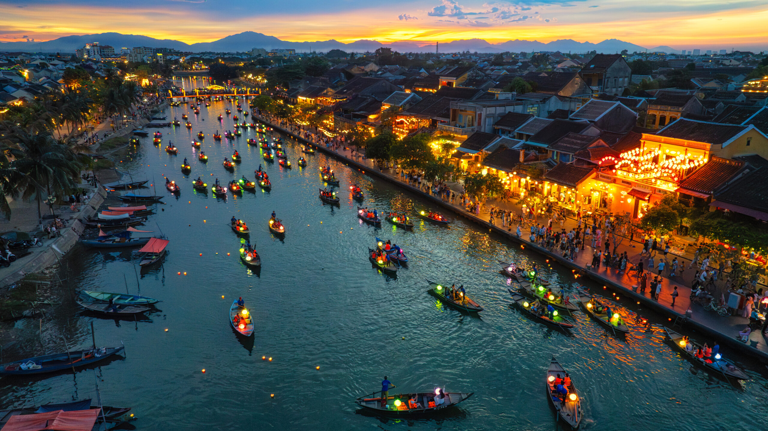 Evening river festival with numerous lit boats along a waterway and crowded riverside streets under a sunset sky.