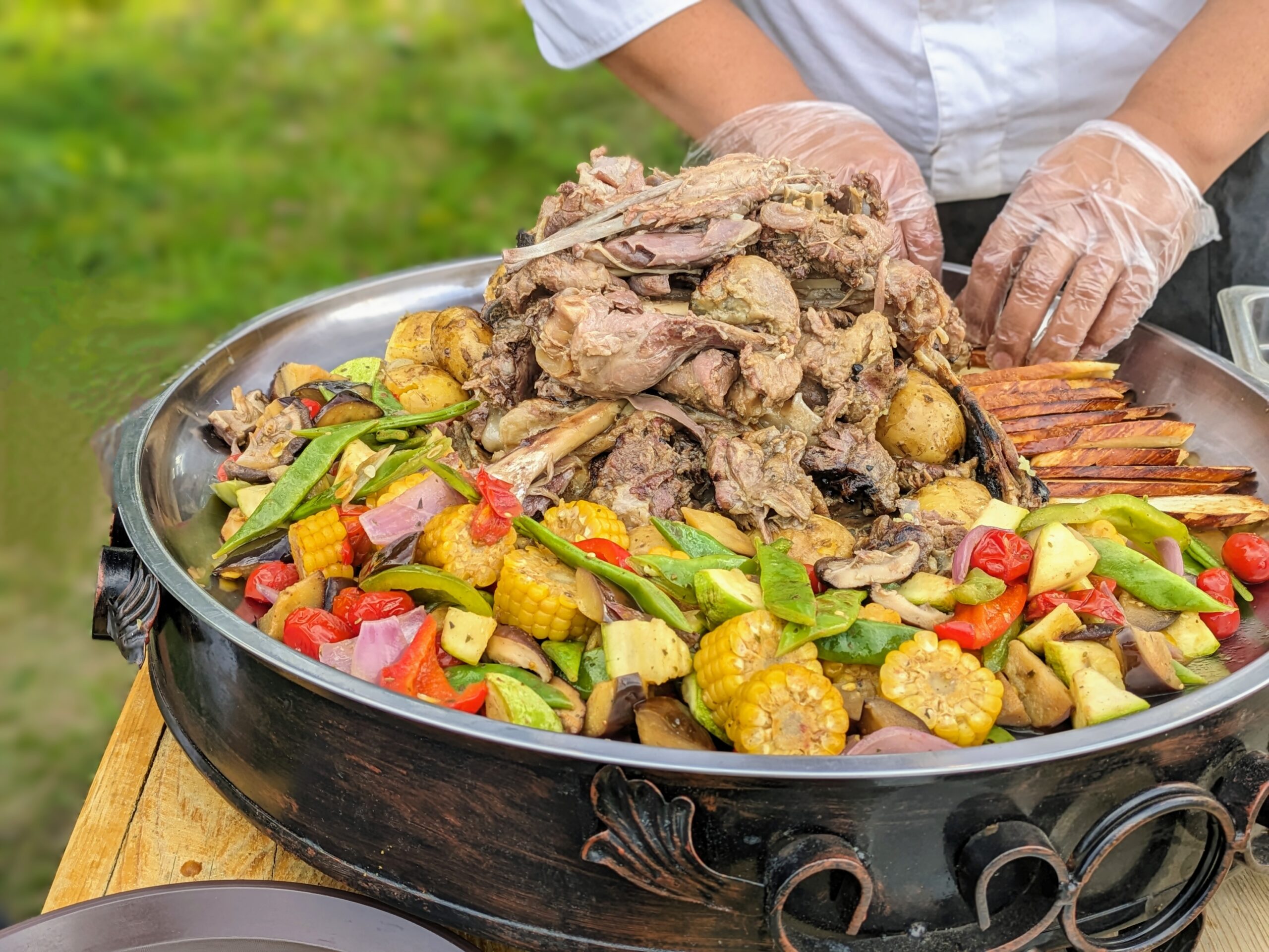 Pile of roasted meat on a large metal platter with corn, peppers and vegetables, being arranged outdoors by gloved hands.