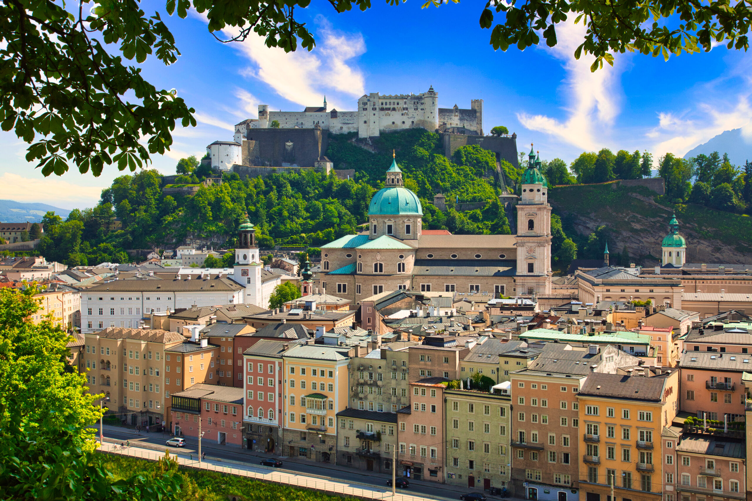 Castle on a hill overlooking a colorful city with green-domed churches and a blue sky, framed by leafy branches.