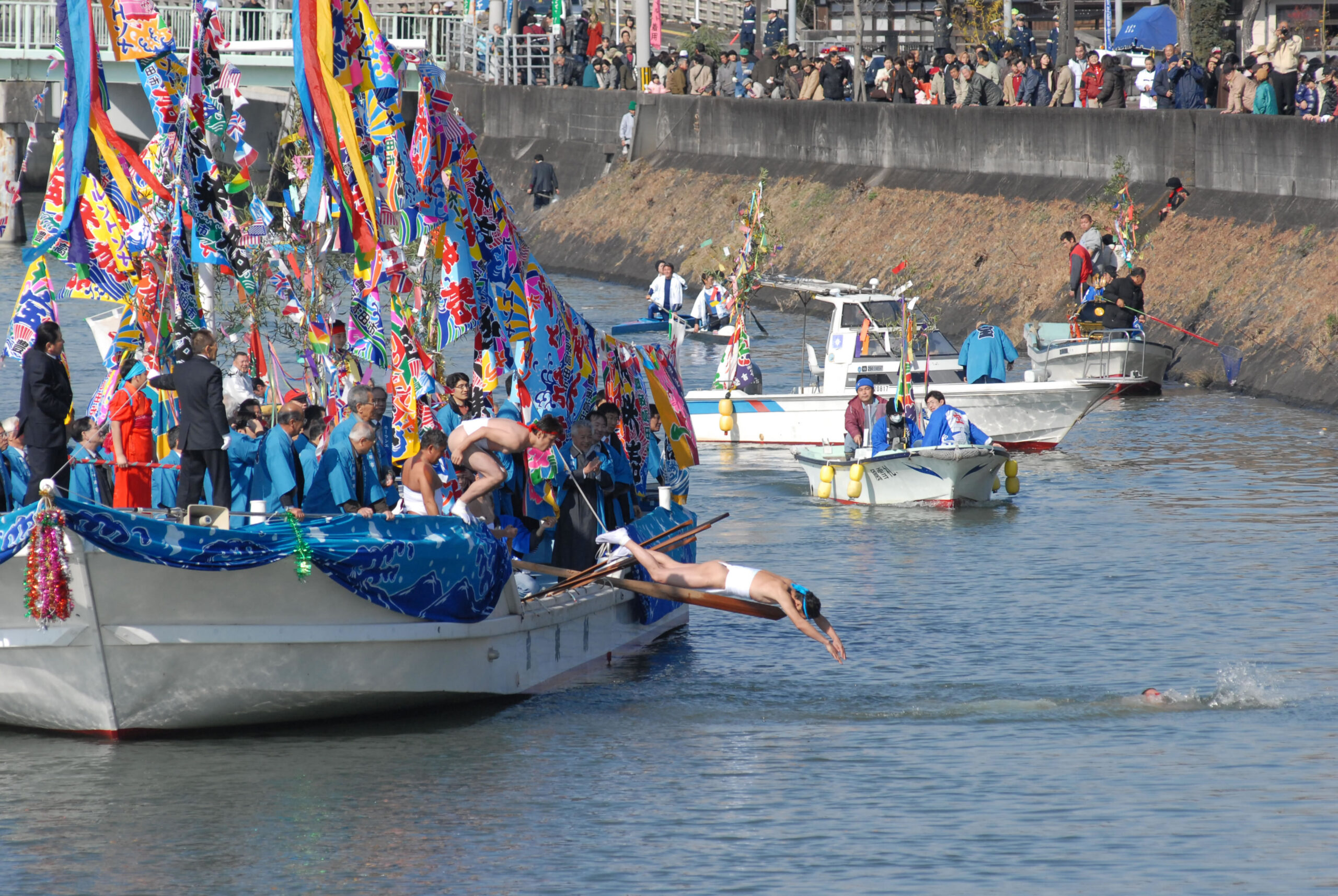 Colorful festival boats with bright flags on a river, spectators line the riverbank watching the event.