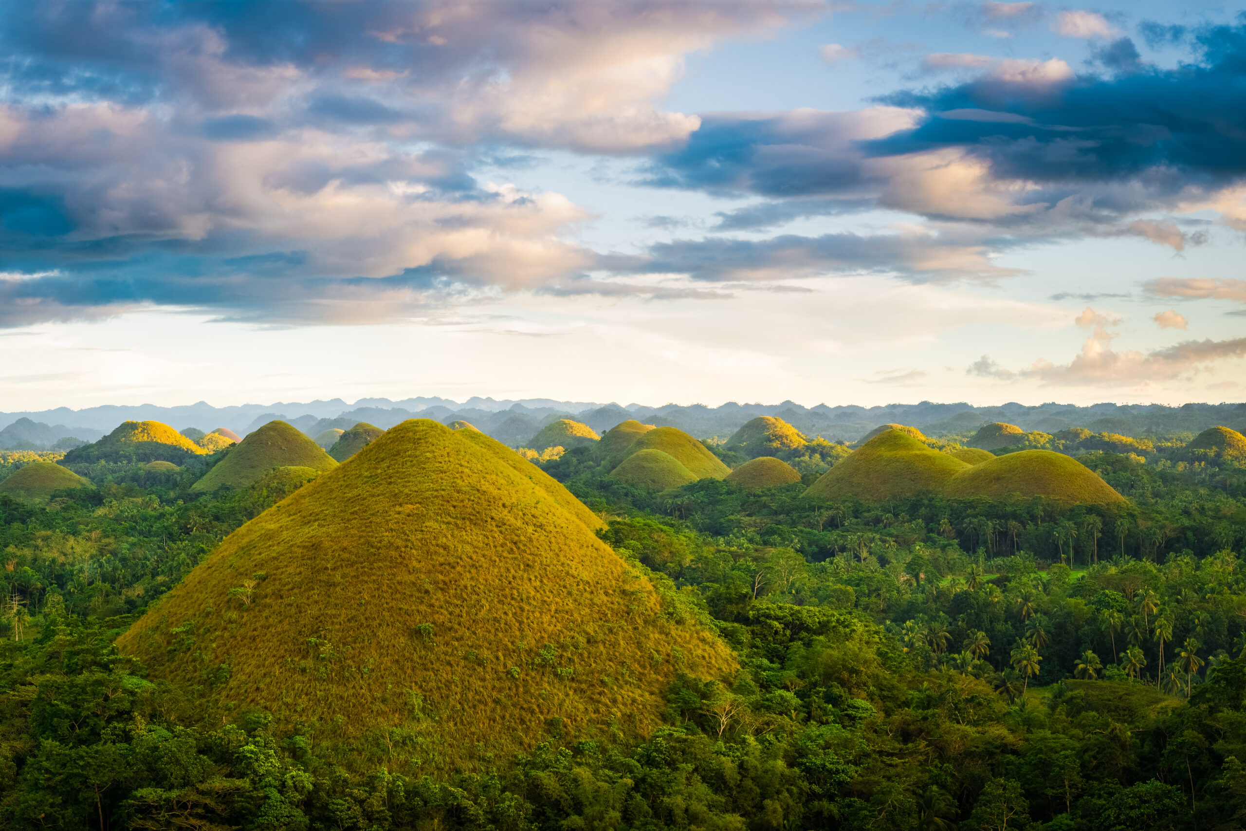 Row of green, dome-shaped hills rising from a dense tropical forest under a partly cloudy sky.