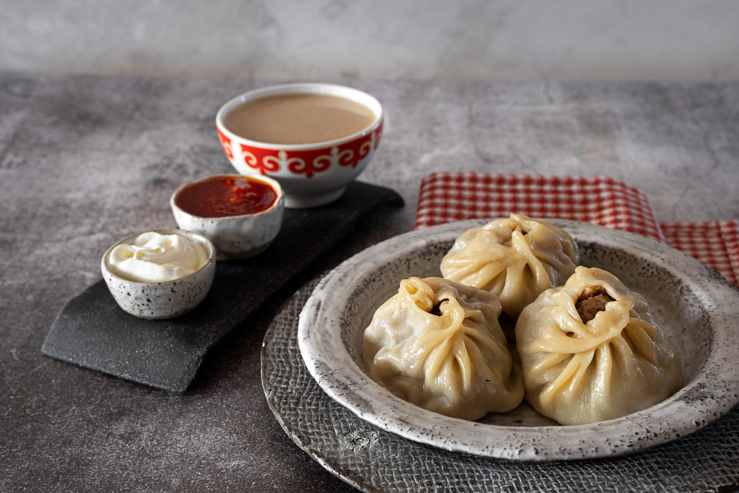 Plate of steamed dumplings with two dipping sauces and a red patterned bowl in the background on a textured surface, inviting and tasty-looking.