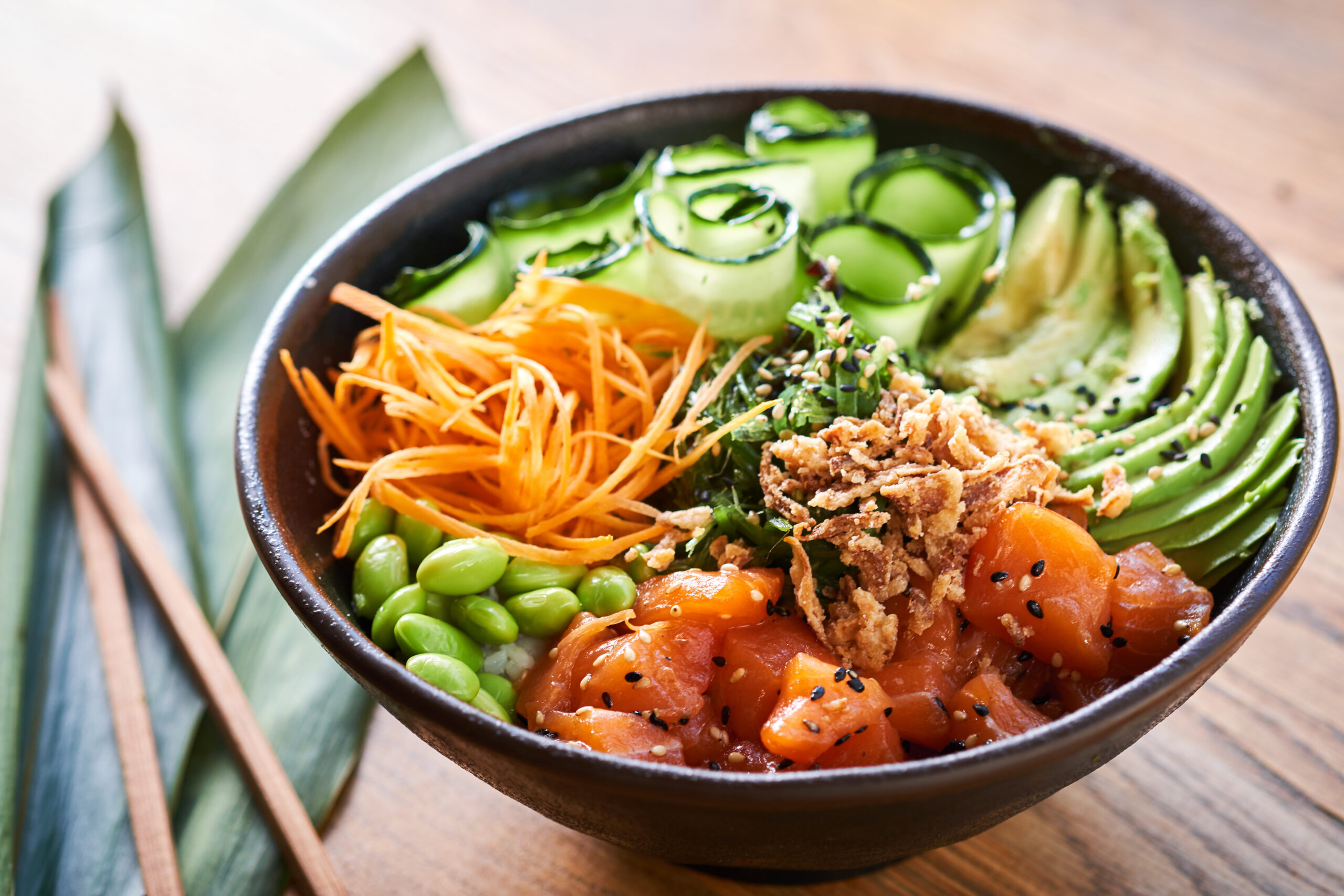 Bowl of poke with salmon, avocado, shredded carrot, edamame, sesame seeds, and crispy onions arranged in a dark bowl on a wooden table
