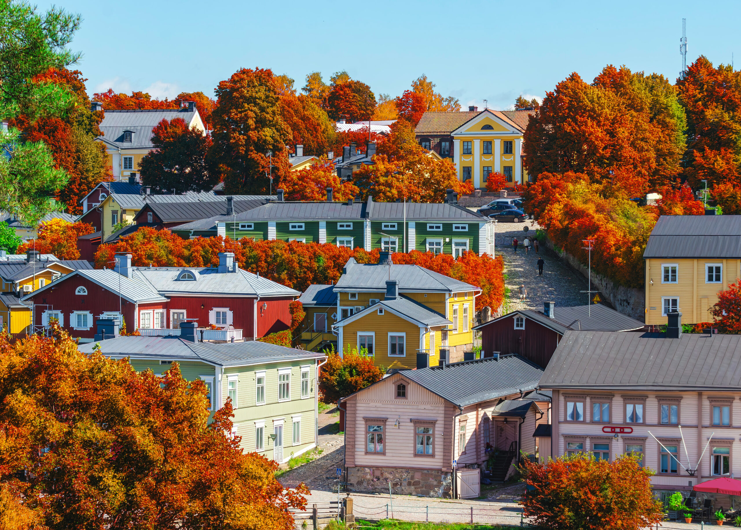 Pastel houses line a cobblestone street as vivid orange-red autumn trees frame a sunny town scene under a blue sky.