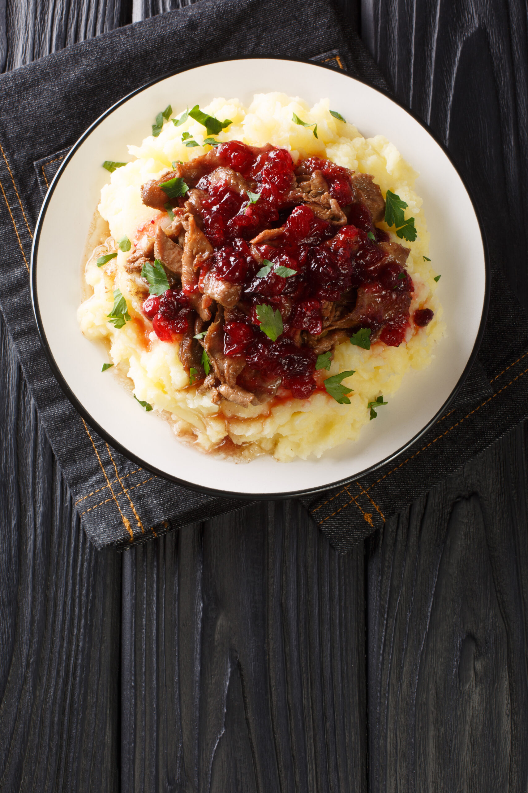 Plate of creamy mashed potatoes topped with braised beef and cranberry sauce, garnished with parsley on a dark wooden table.
