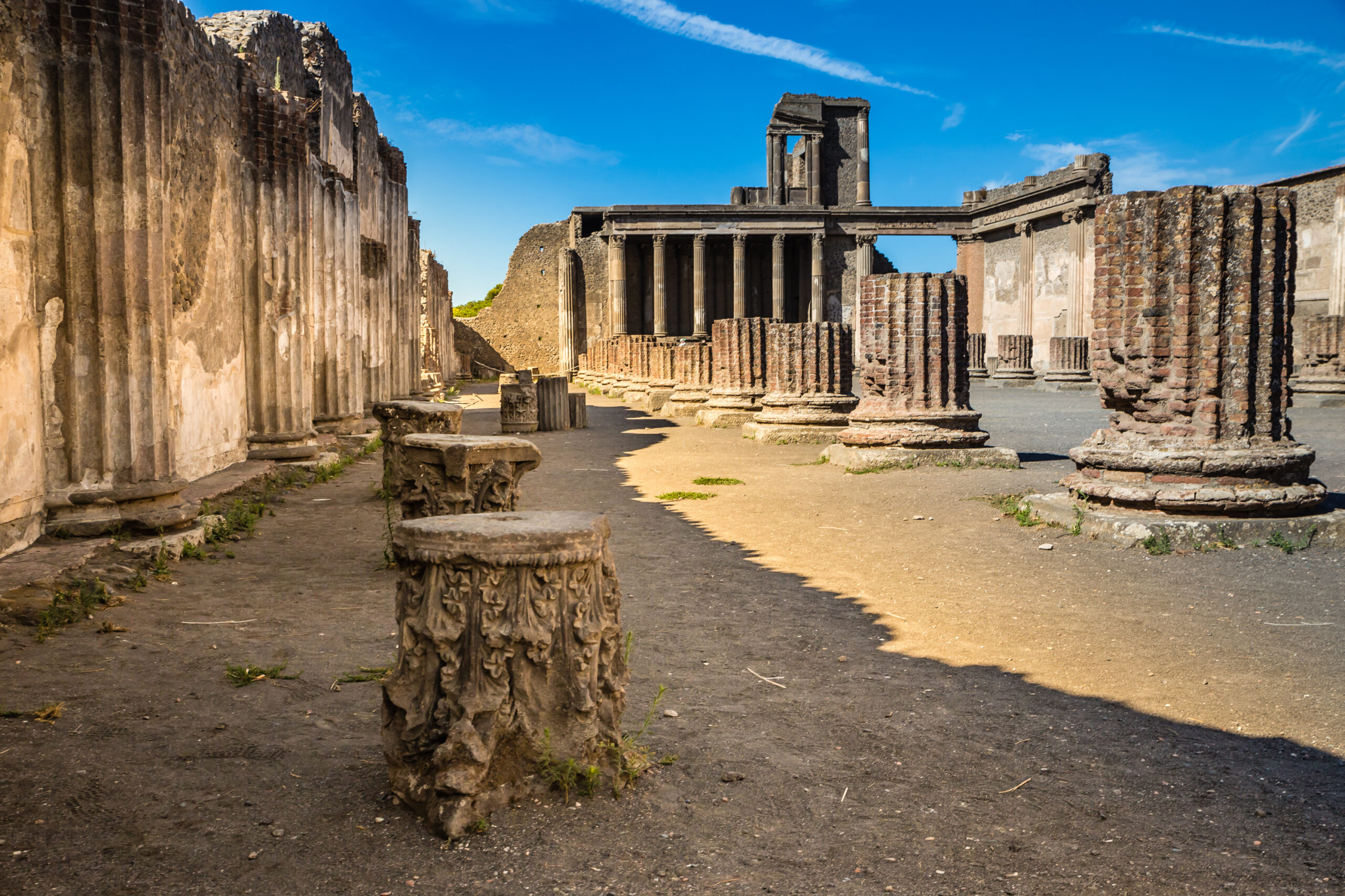 Row of ancient stone columns forming a sunlit colonnade in a ruinous archaeological site against a bright blue sky, with scattered carved stone bases in the foreground.