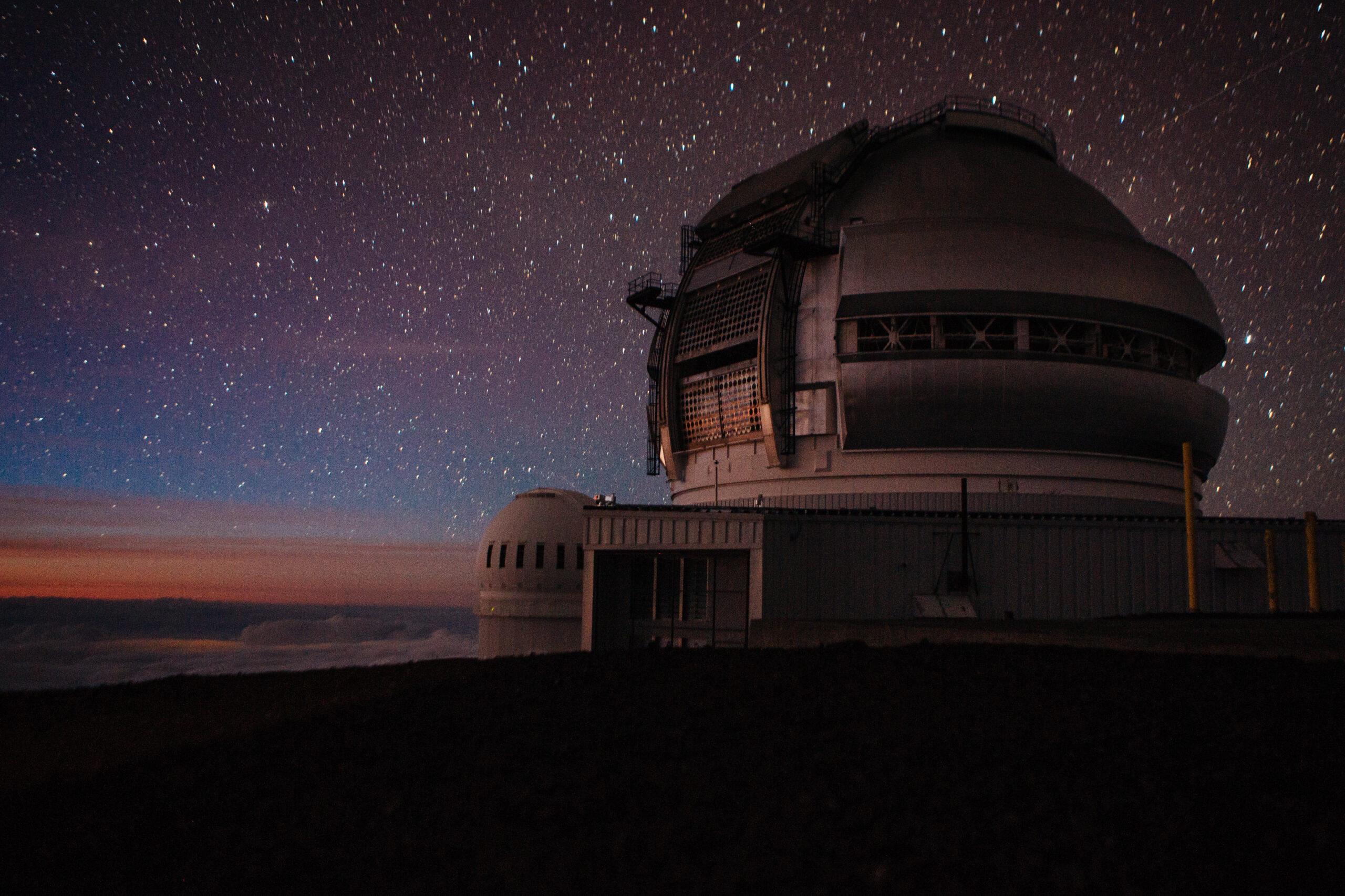 Night sky filled with stars above a large observatory dome at dusk with a railing along the top and dark landscape in the foreground.