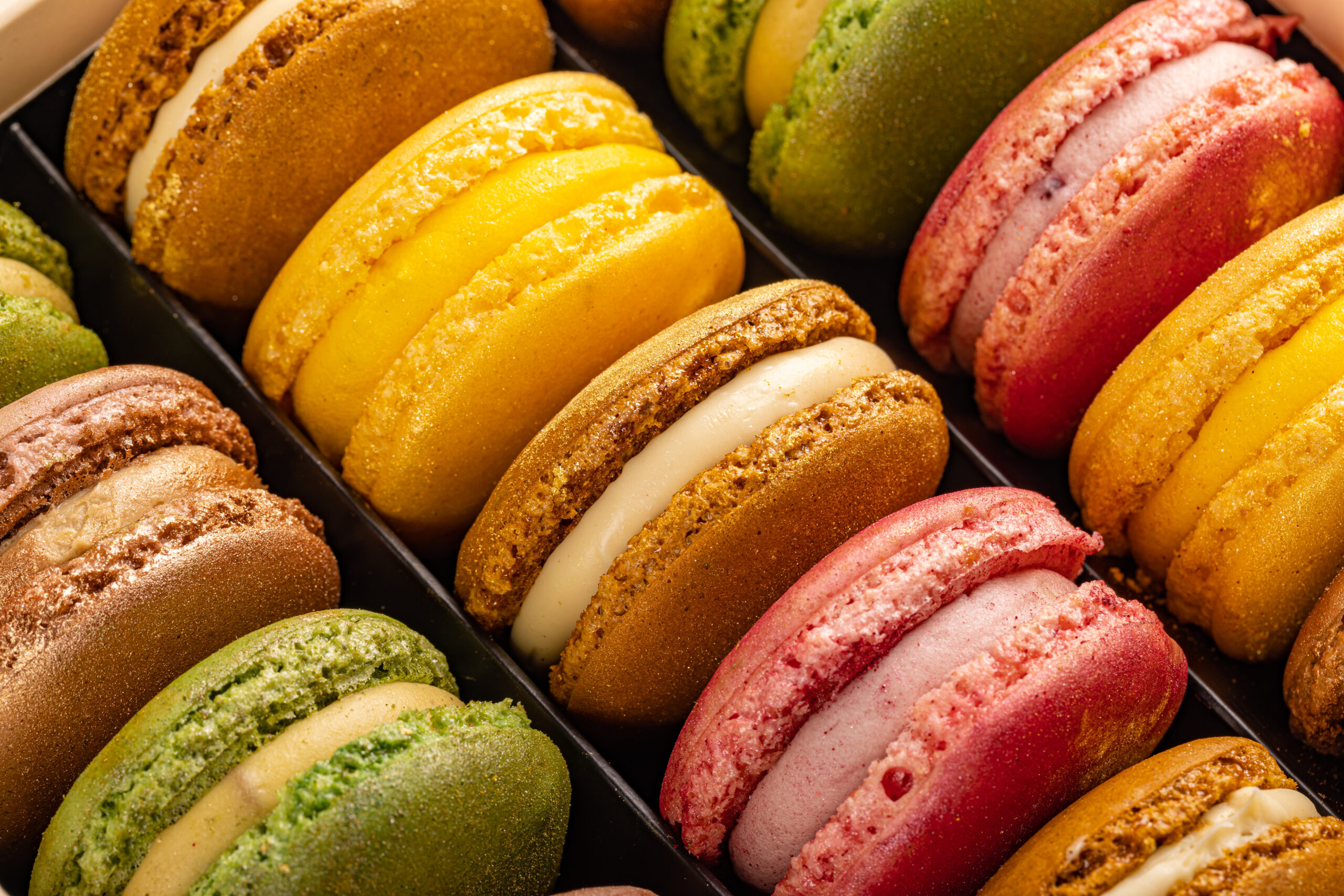 Assorted colorful macarons in a bakery box, with green, yellow, pink, and brown shells and cream fillings.