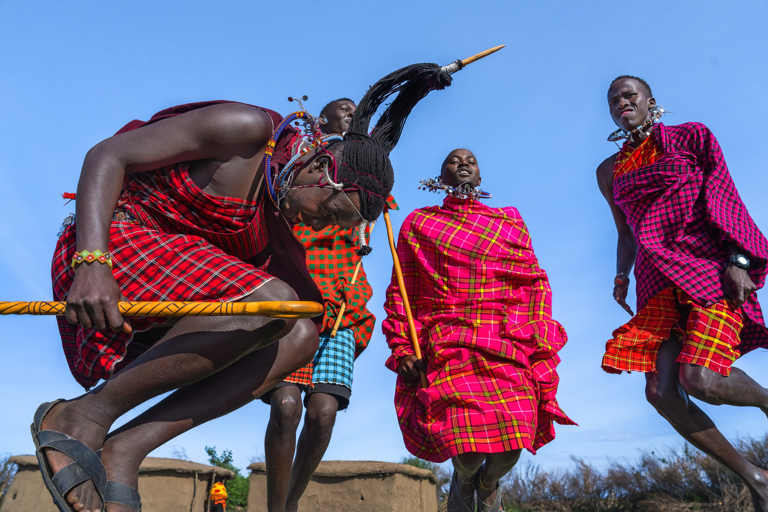 Maasai people in red plaid shukas performing a jumping dance against a clear blue sky.