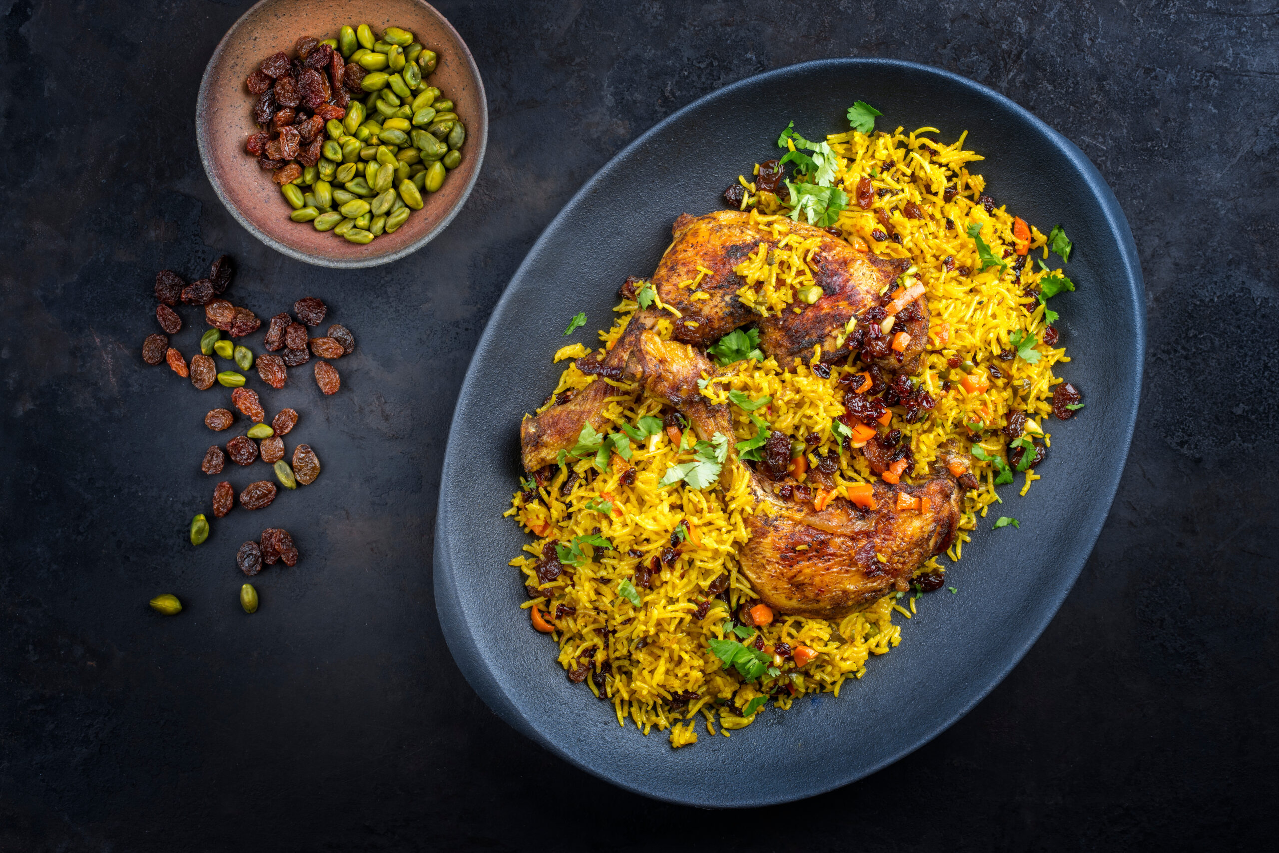 Plate of yellow rice with seasoned chicken, garnished with herbs, and scattered raisins and pistachios on a dark platter.