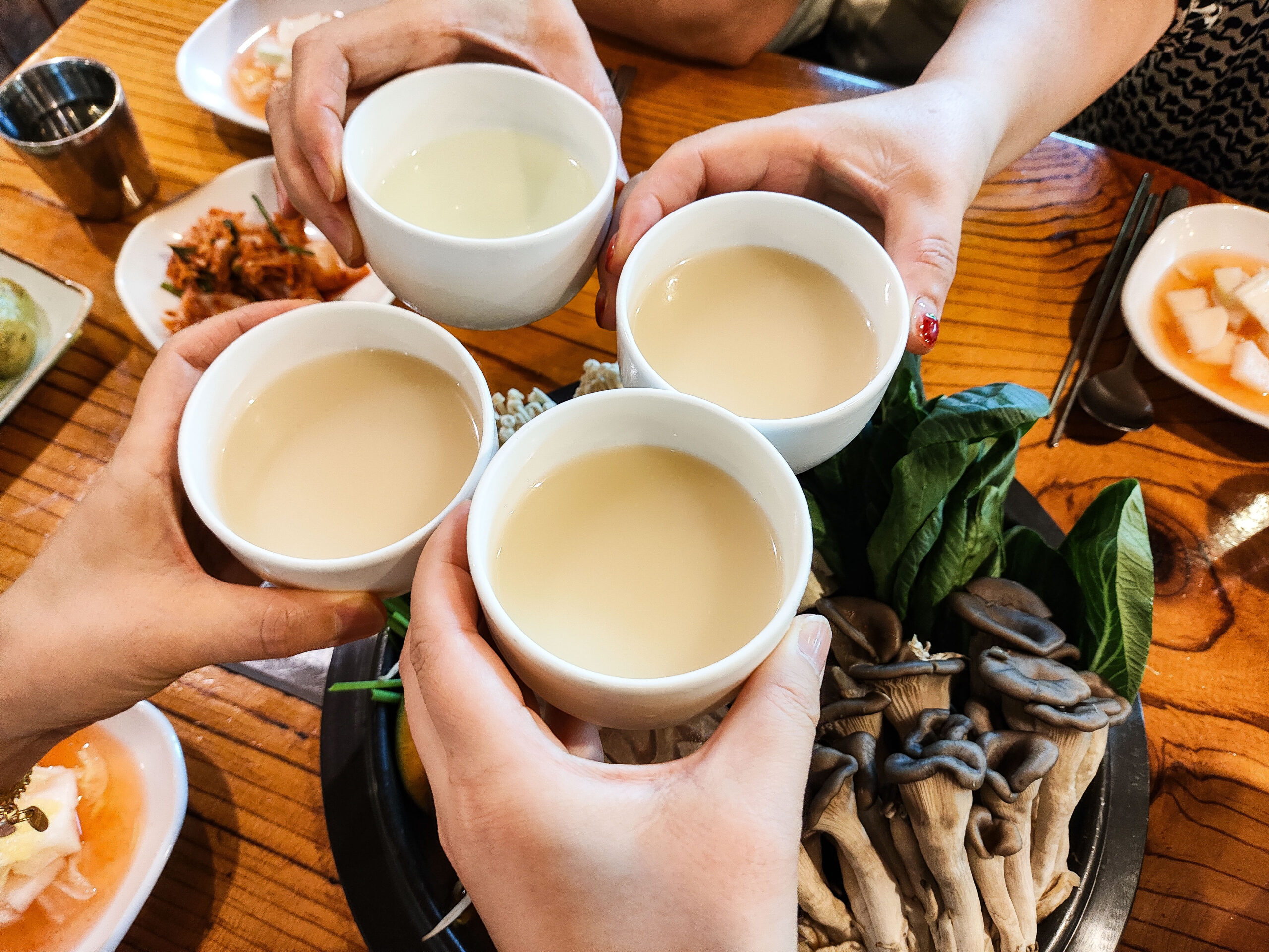Peers raise white cups in a toast over a wooden table with plates of food nearby.