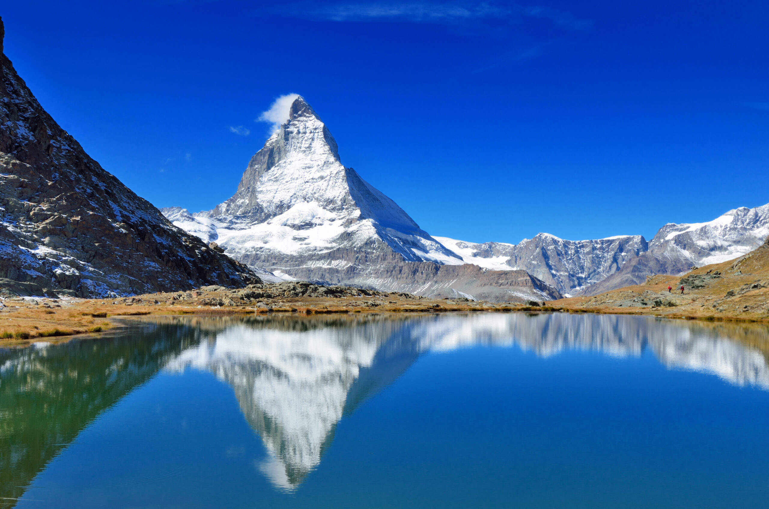 Snow-capped Matterhorn reflected in a calm alpine lake, with rocky foreground and distant hikers on the right under a clear blue sky.