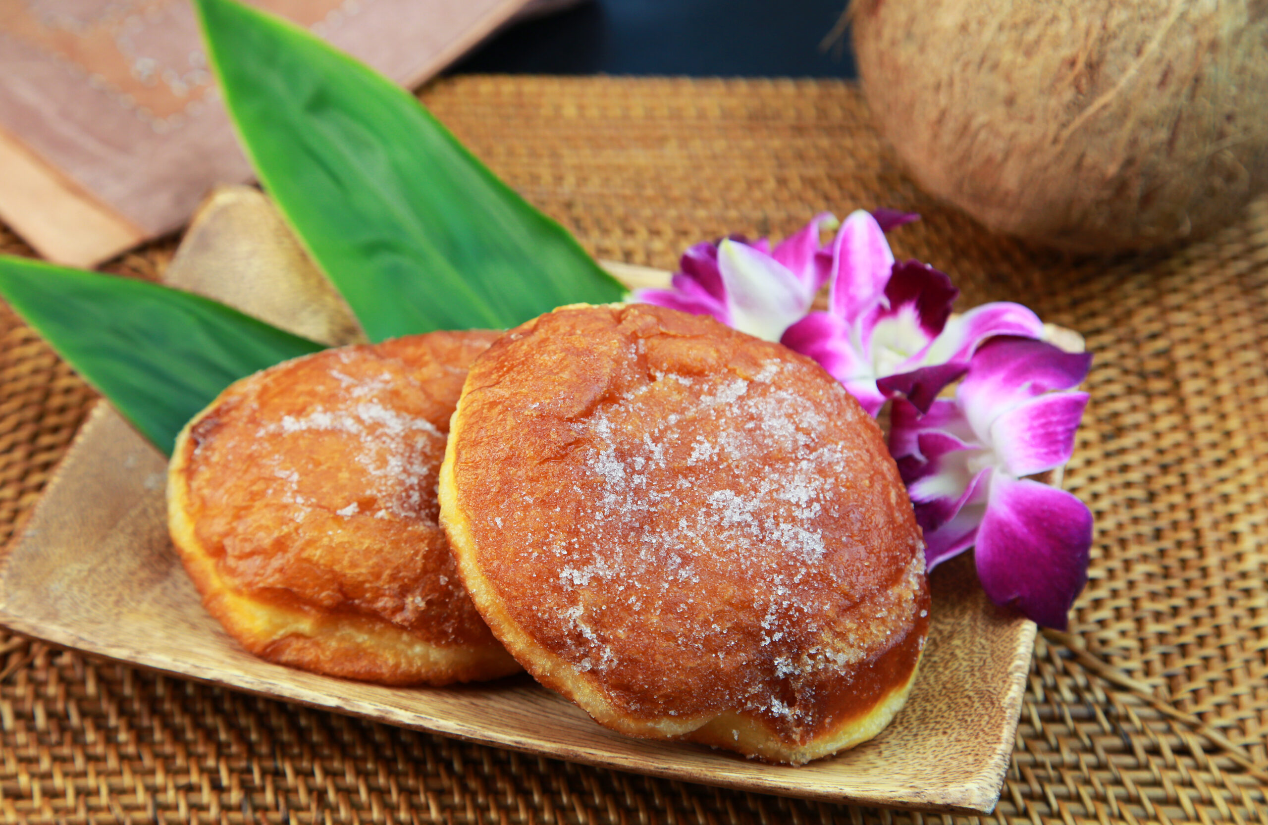 Three sugar-dusted donuts on a wooden tray with green leaves and a purple orchid in the background.