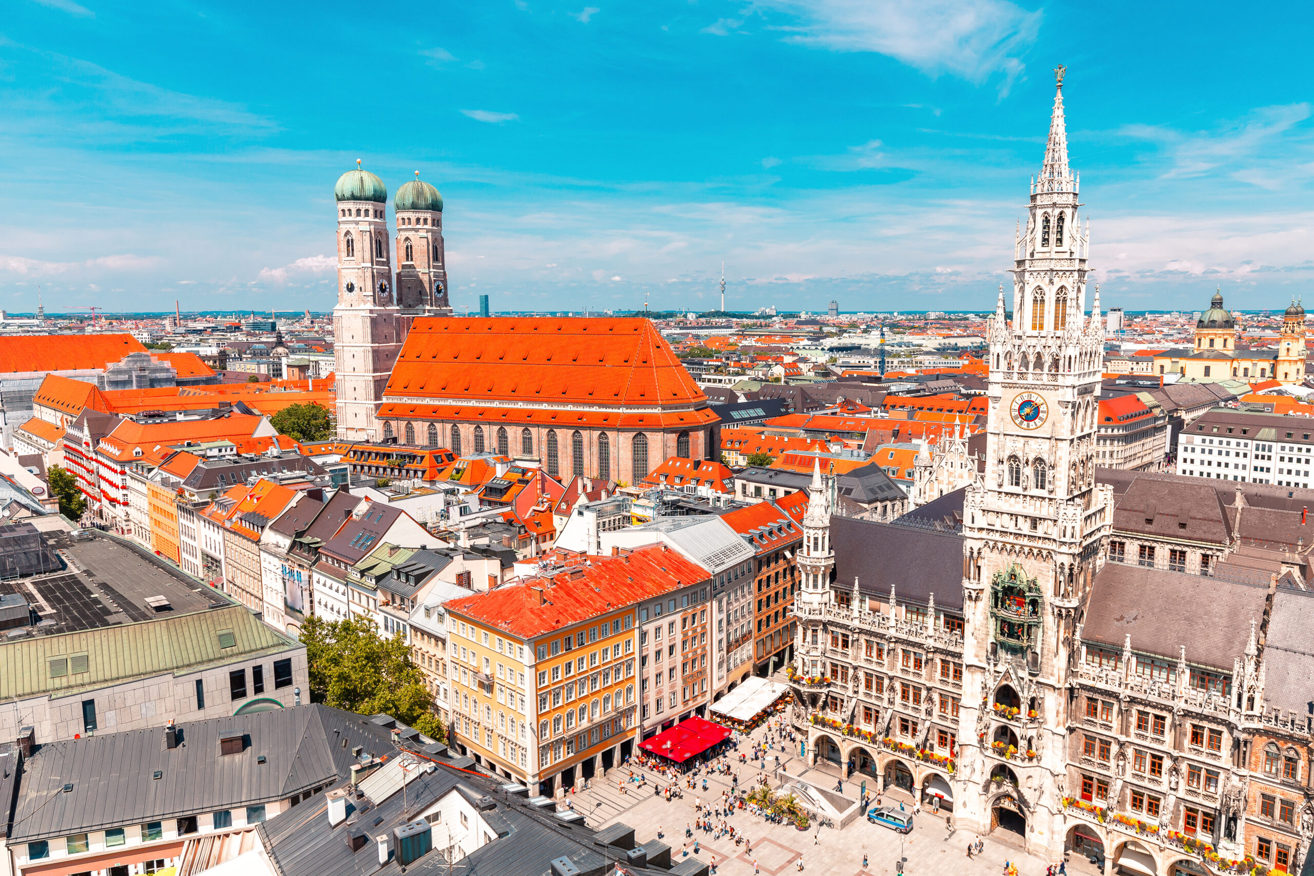 Aerial view of Munich city center with red rooftops, the Frauenkirche with green domes, and the white New Town Hall against a blue sky.