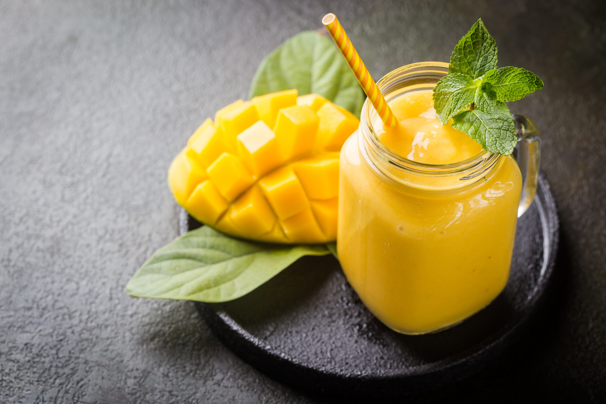 Mango smoothie in a mason jar with a striped yellow straw and fresh mint, beside diced mango on a dark surface.