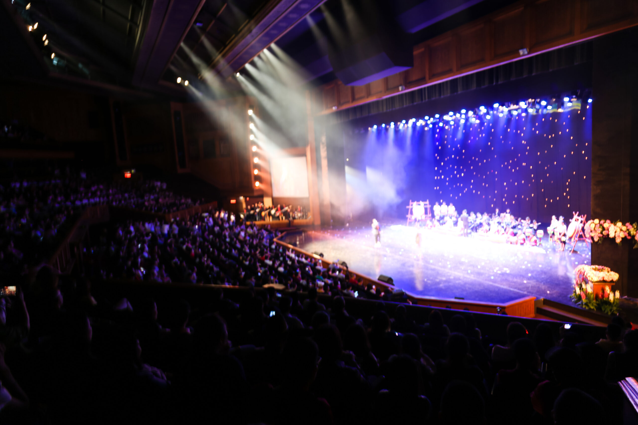Audience watching a live stage performance in a large theater with bright blue backdrop and stage lights illuminating performers.