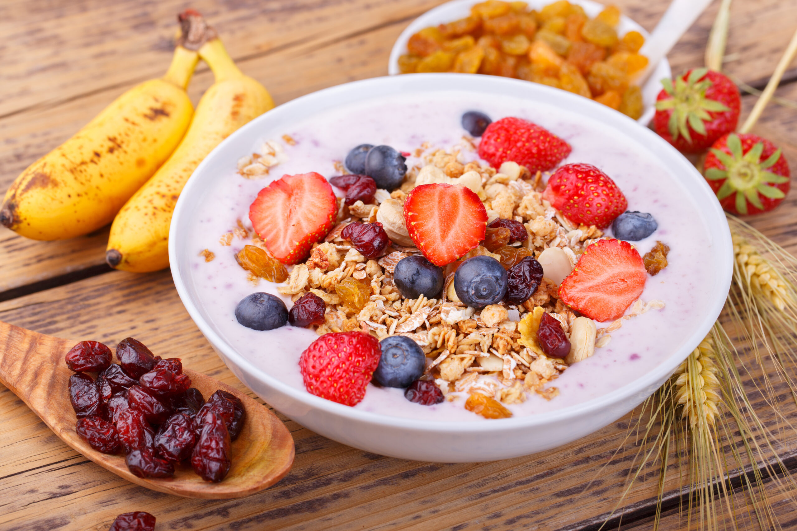 Creamy yogurt parfait topped with sliced strawberries, blueberries, granola and raisins in a white bowl on a wooden table, with bananas nearby.