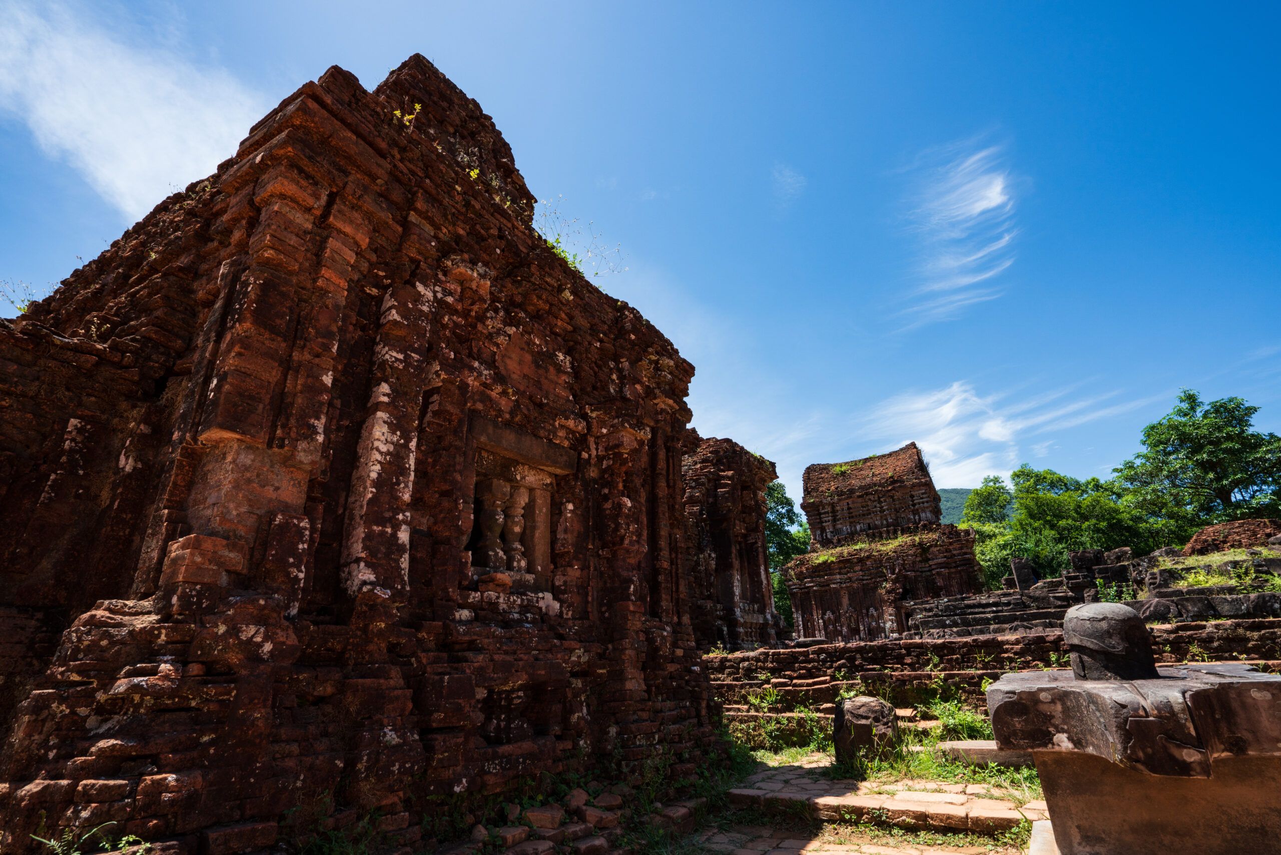 Ruined red-brick walls rise at an angle, with stacked rectangular blocks and overgrown plants, under a clear blue sky.