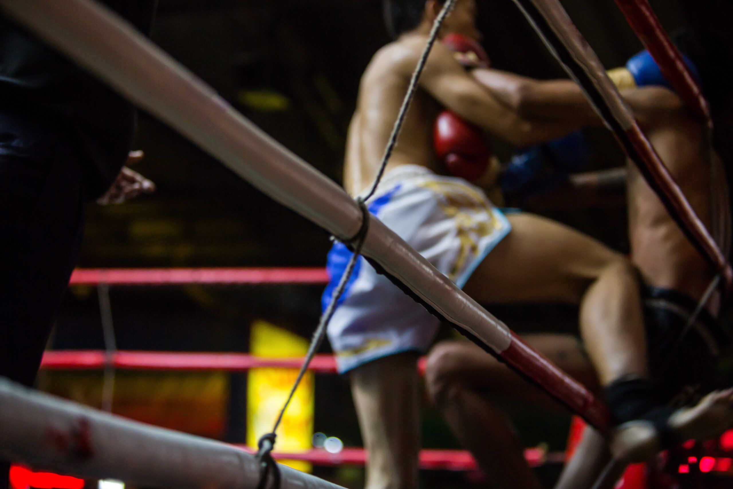 Boxer in white shorts with red gloves inside a boxing ring, ropes crossing the foreground and dim arena lights in the background.