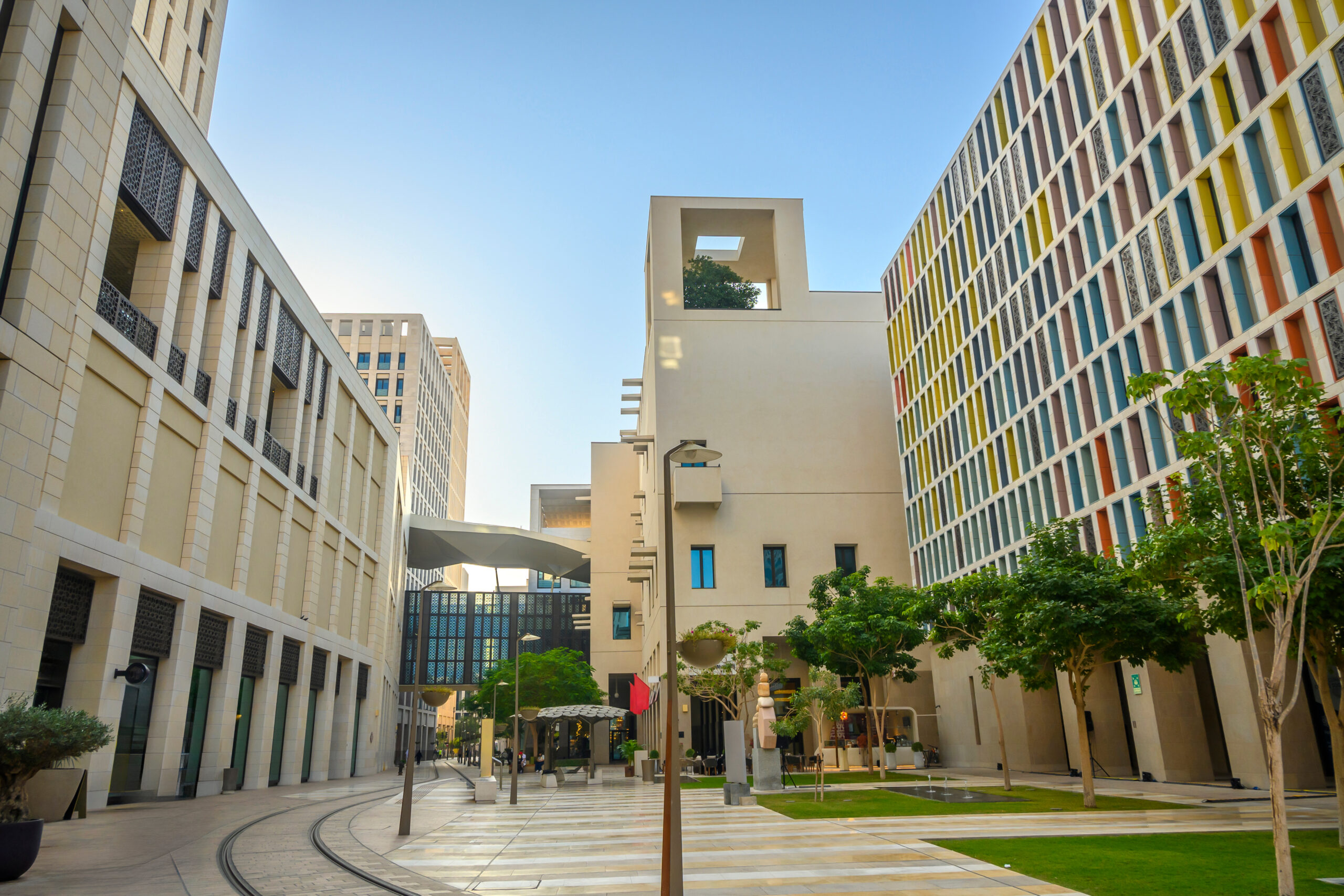 Wide view of a modern open-air courtyard between beige buildings, with a colorful façade on the right and trees against a clear blue sky.
