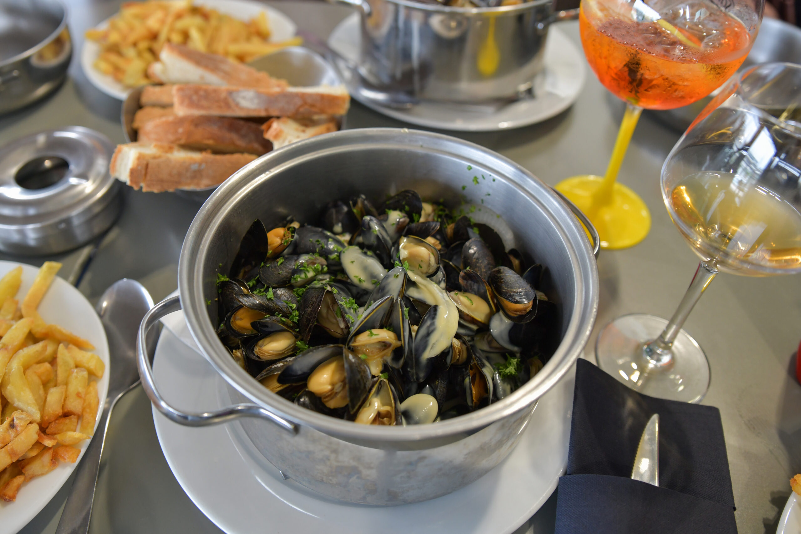 Mussels in a metal pot with cream sauce and parsley on a table surrounded by bread, fries, and wine glasses