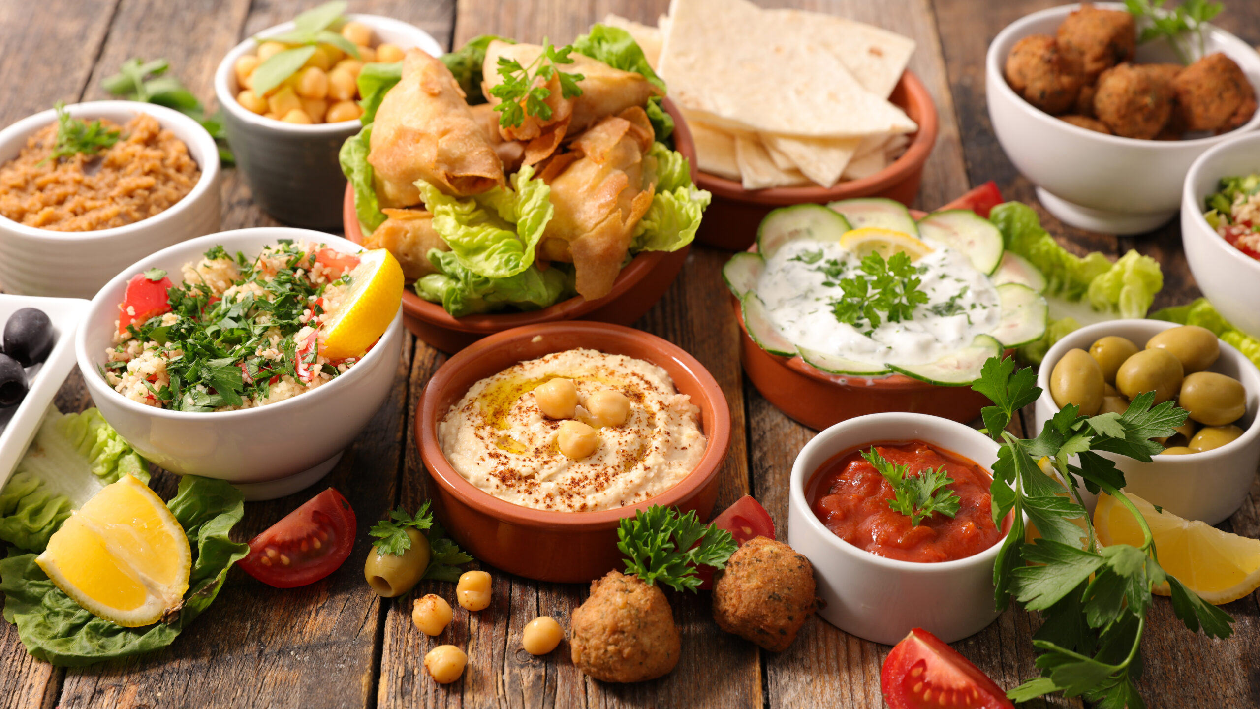 Assorted Mediterranean tapas on a rustic wooden table, including hummus, olives, chickpea salad, cucumber yogurt dip, pita, falafel, and lemon wedges.