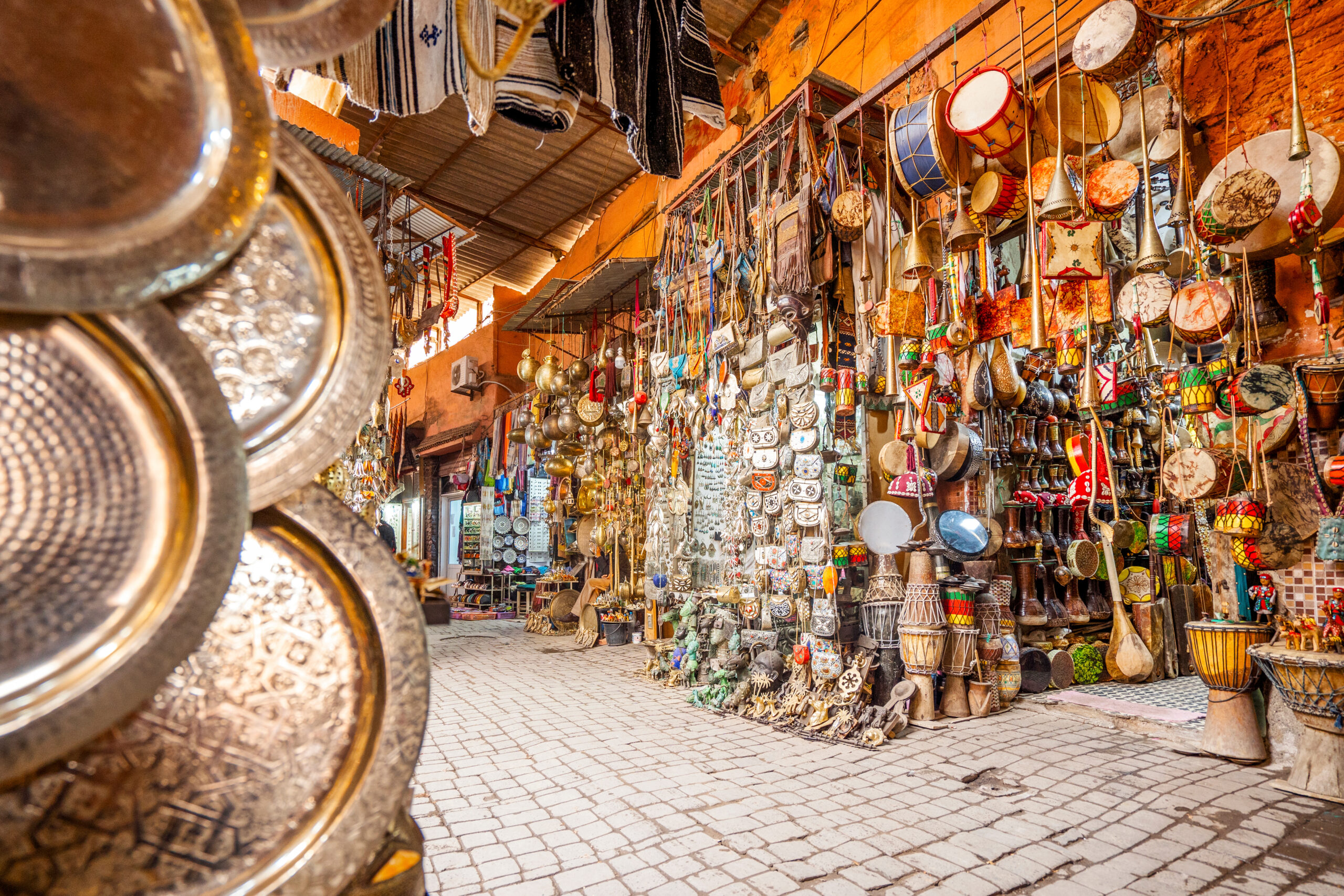 Bustling market stall with hanging brassware, drums, and pots in vibrant colors under a wooden awning