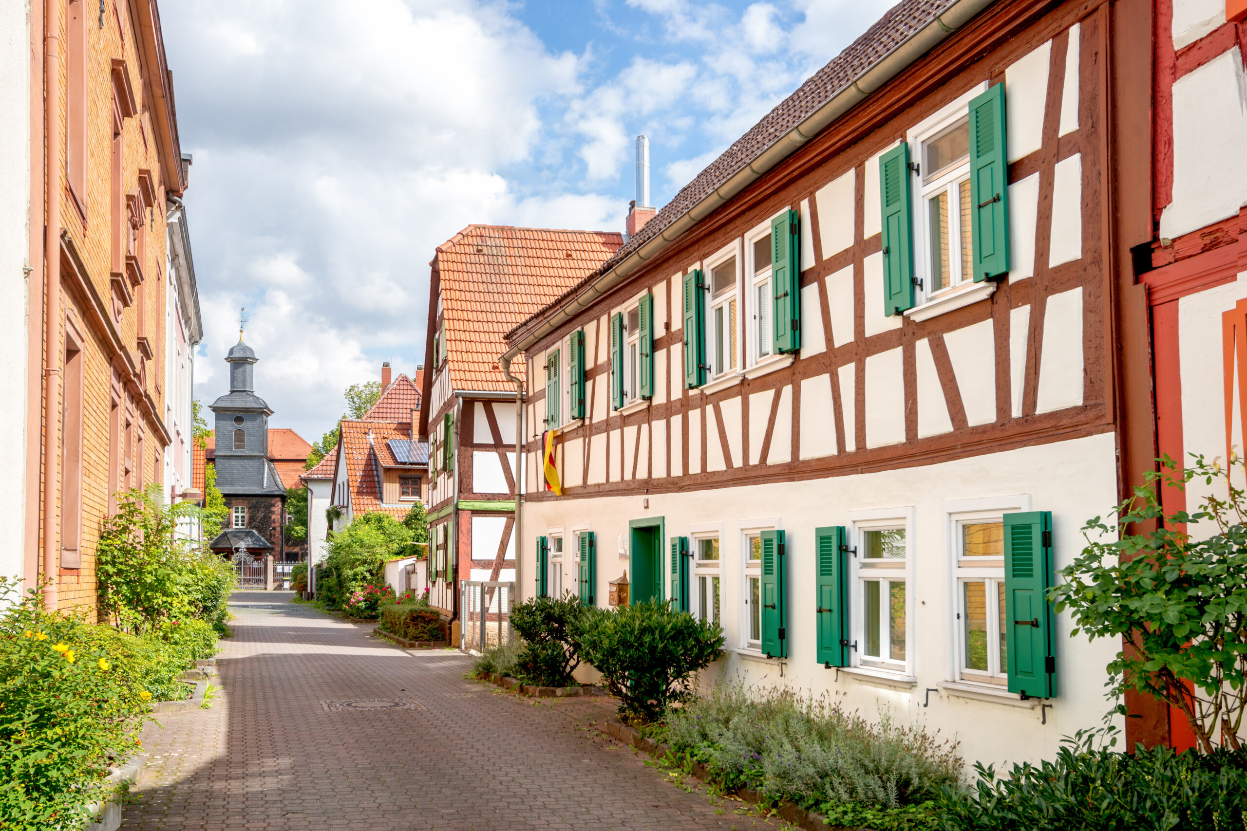 Colorful European street scene with half-timbered white walls, brown timber, green shutters, and red-tiled roofs under a blue sky.